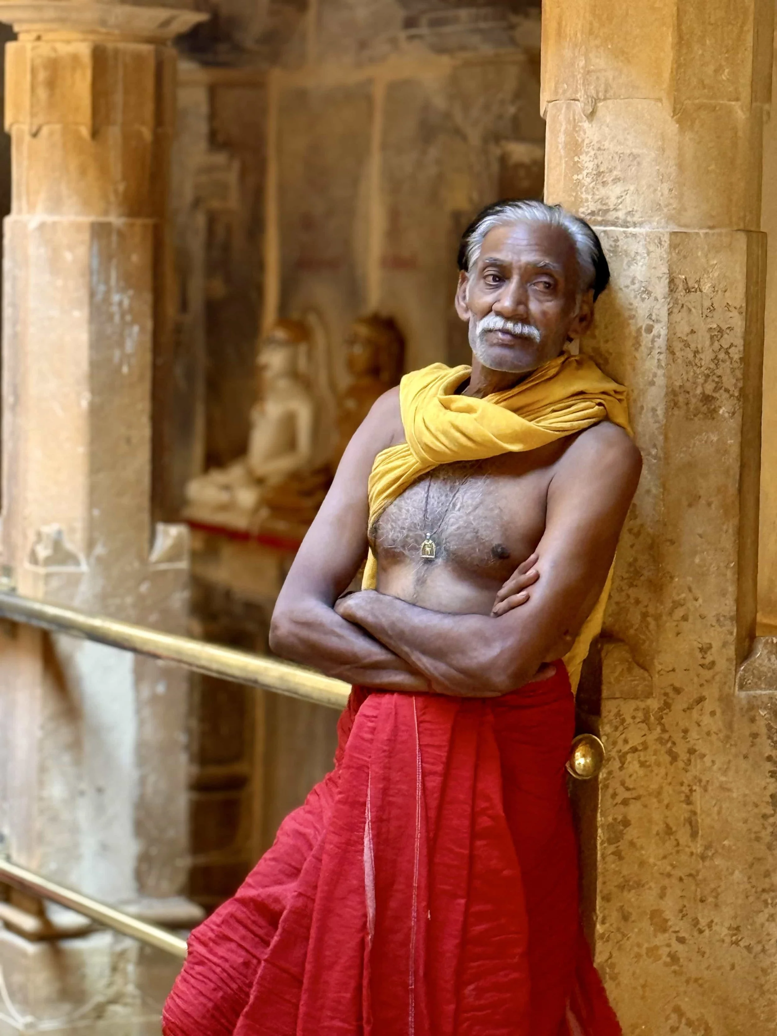 An elderly man with gray hair and a mustache leaning against a stone wall inside a building, wearing a yellowcloth draped over his shoulders and red pants.