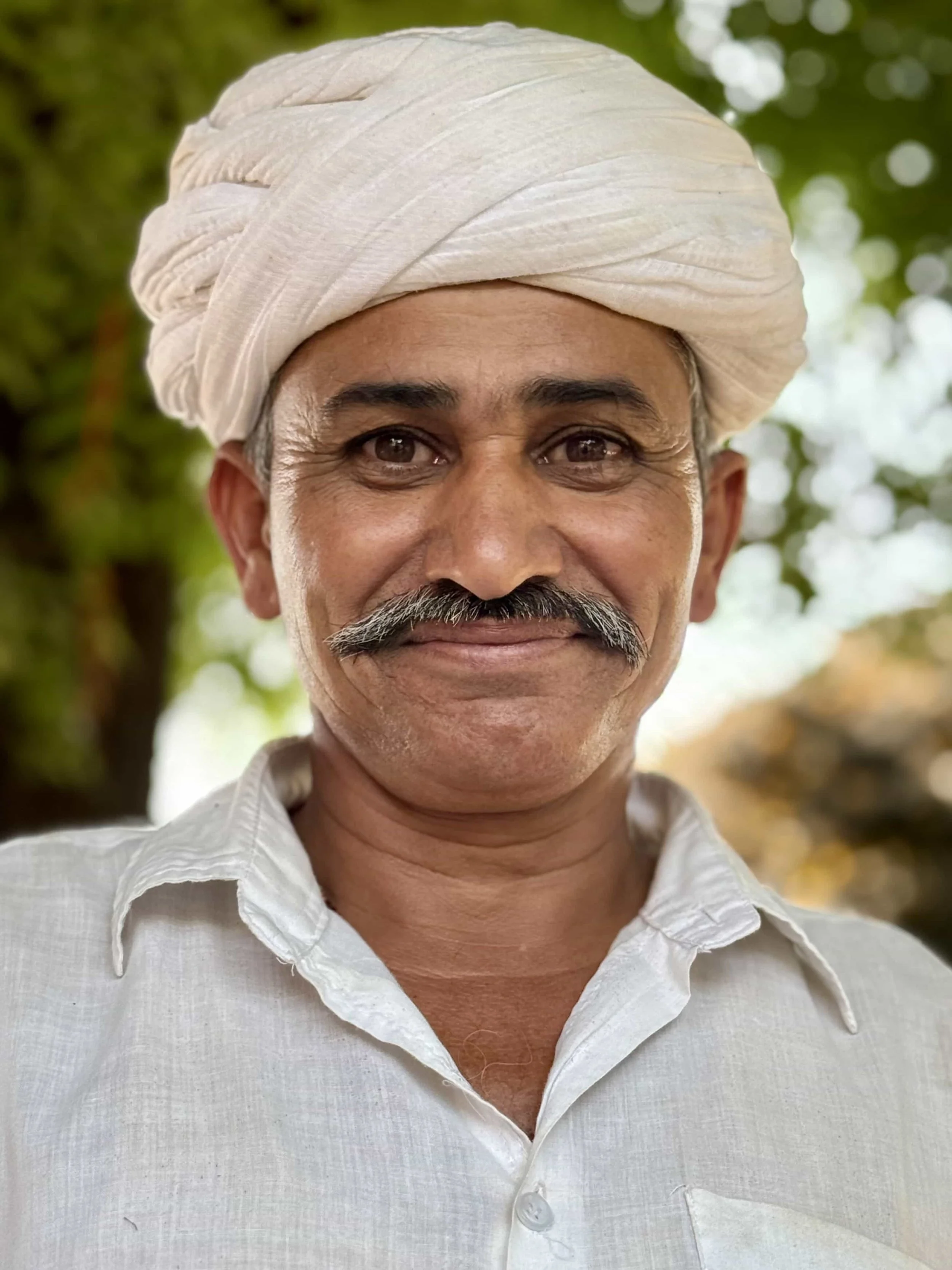 Close-up of a smiling middle-aged man wearing a white turban and white shirt, outdoors with blurred green and yellow foliage in the background.