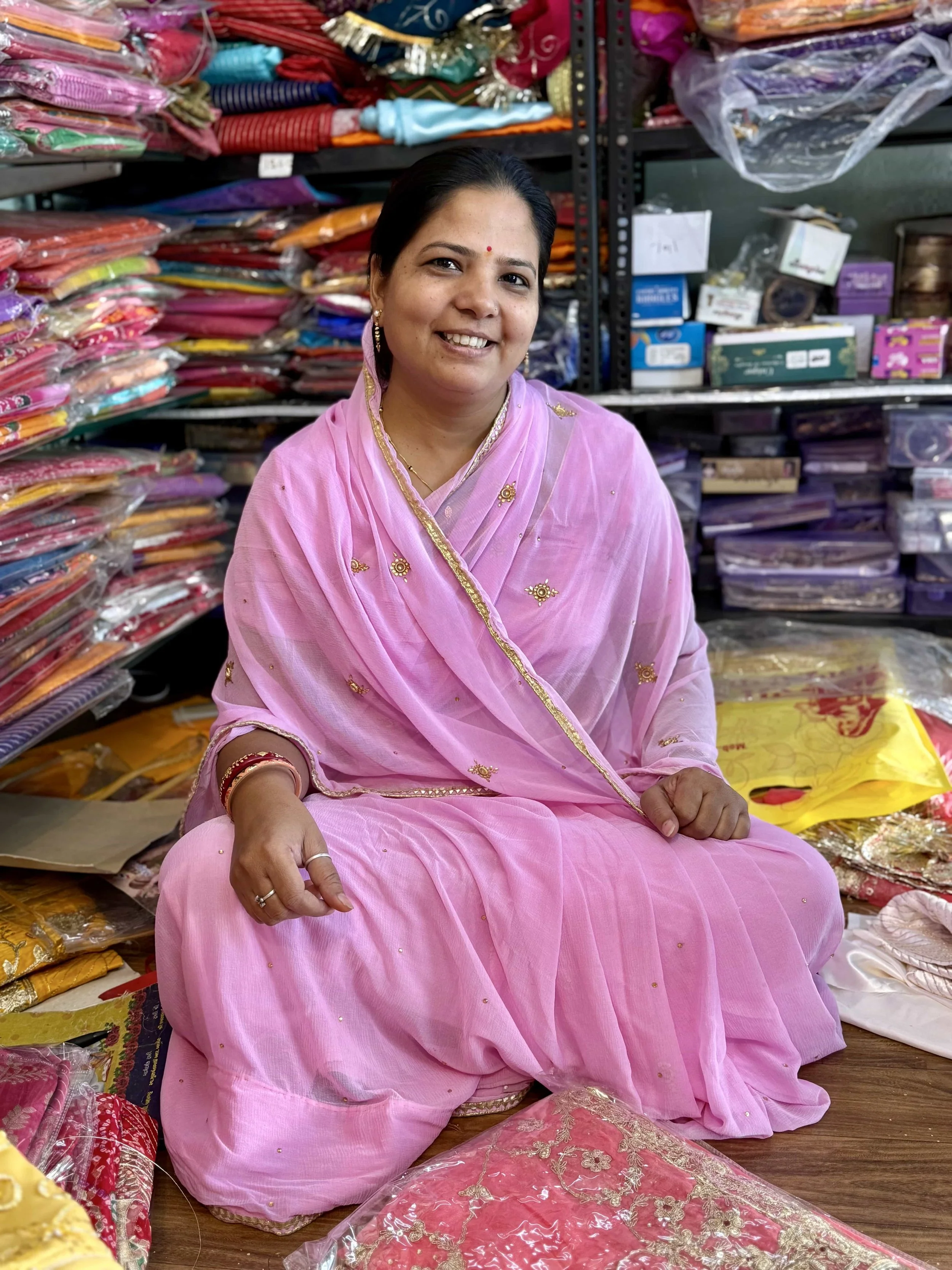 A woman dressed in a pink traditional Indian attire, sitting on the floor in a shop surrounded by colorful fabrics and embroidered textiles.