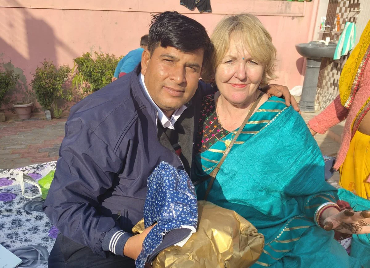 A man and a woman sitting close together outdoors, smiling at the camera. The man has dark hair and is wearing a dark jacket, while the woman has blonde hair and is dressed in traditional Indian attire. The background shows potted plants and a pink wall.