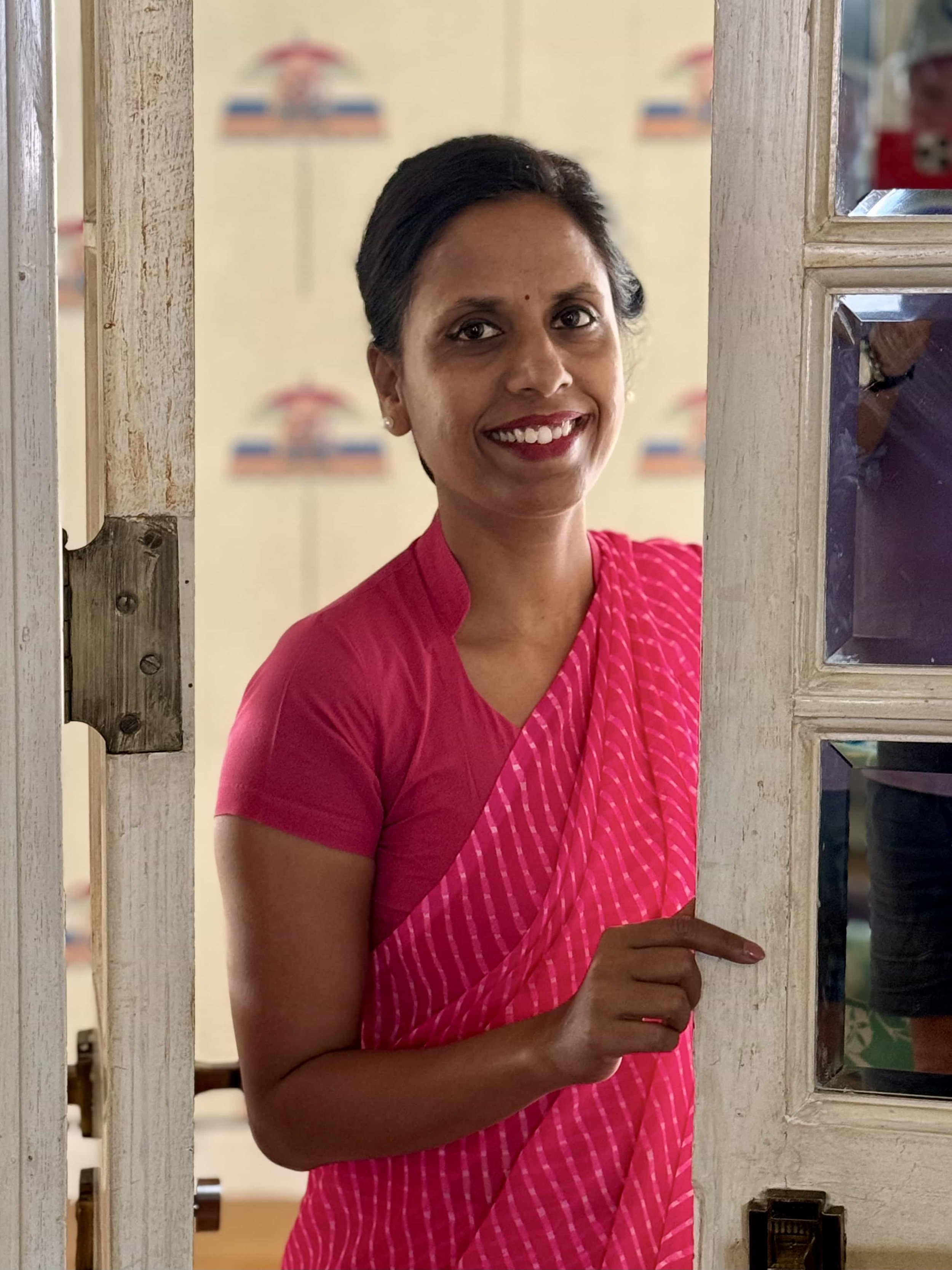 A woman with dark hair smiling, wearing a pink saree with pink and white stripes, standing by a glass-paneled door.