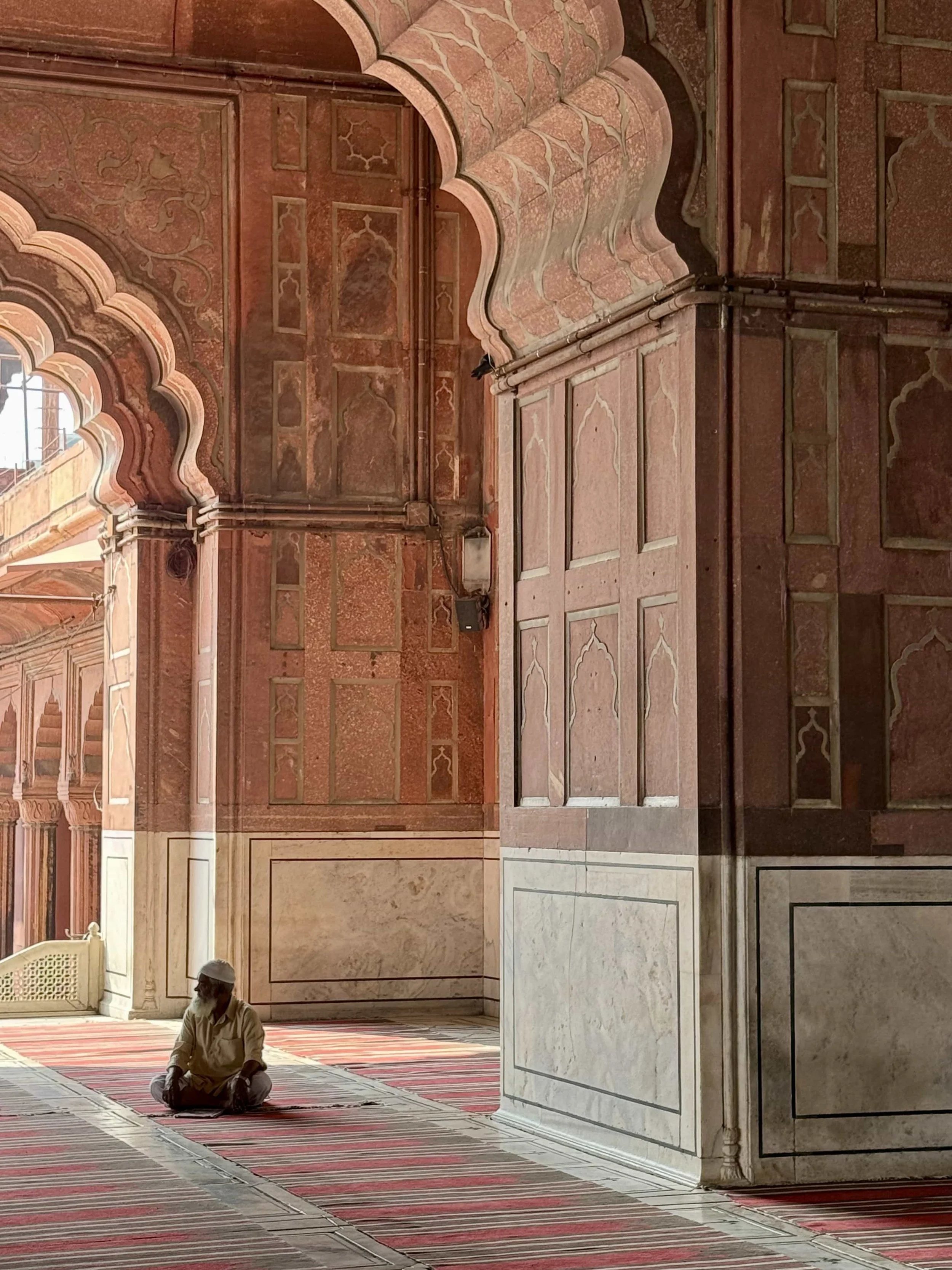 Interior of a mosque with a man sitting on the carpet, intricate wall and ceiling designs, arches, and a window letting in natural light.