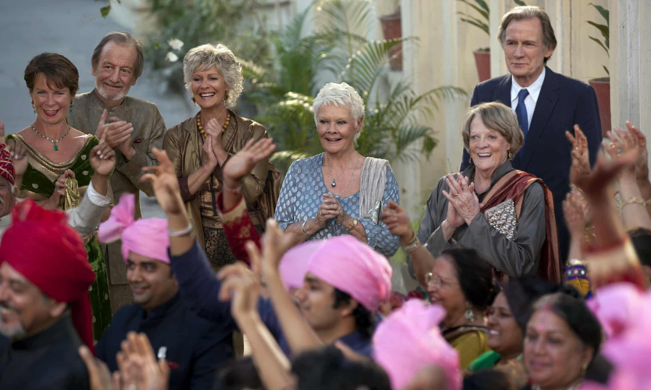 A group of elderly women and men at a celebration, some wearing formal attire, clapping and smiling amidst colorful decorations and pink hats.