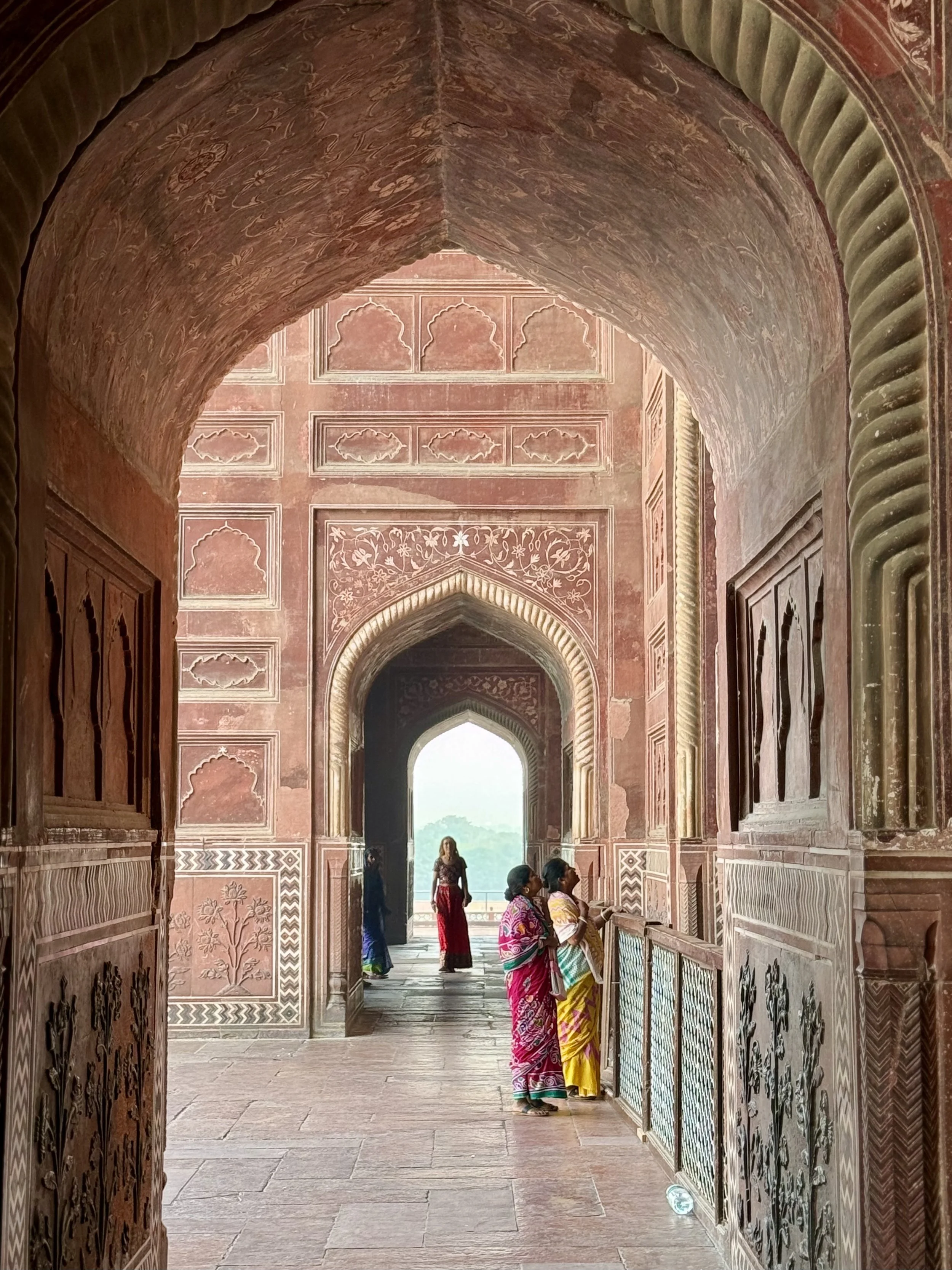 People standing and walking in a decorative hallway with intricate wall and ceiling carvings, framed arches, and natural light coming through an opening at the end.