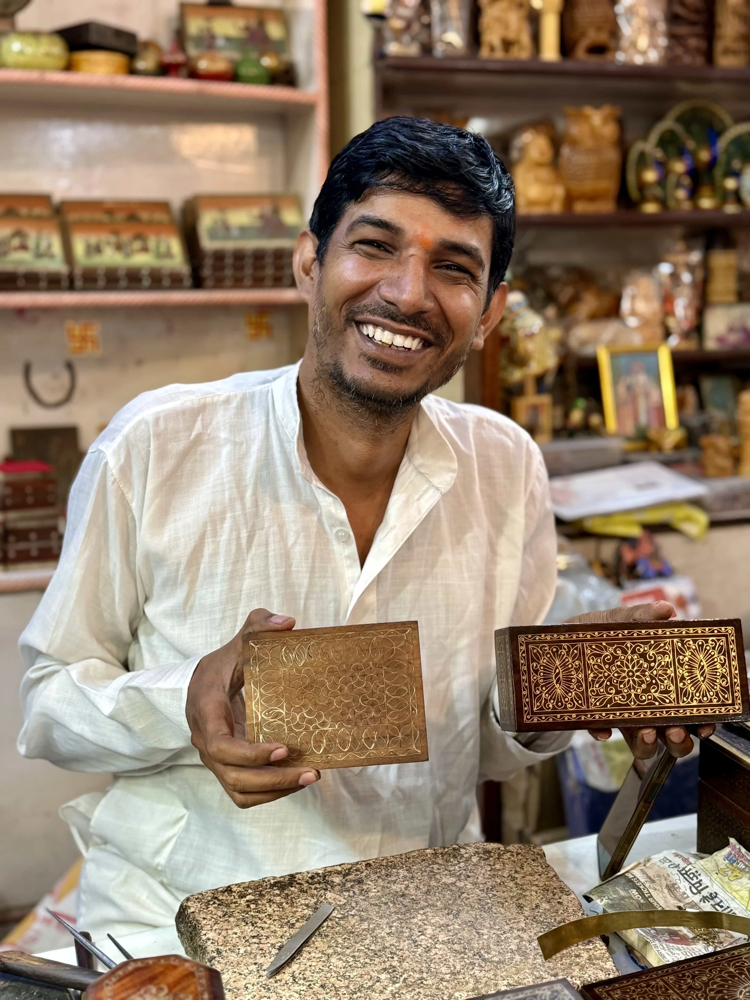 A smiling man with dark hair and a light-colored shirt holding decorative wooden boxes with intricate carvings in a craft shop.