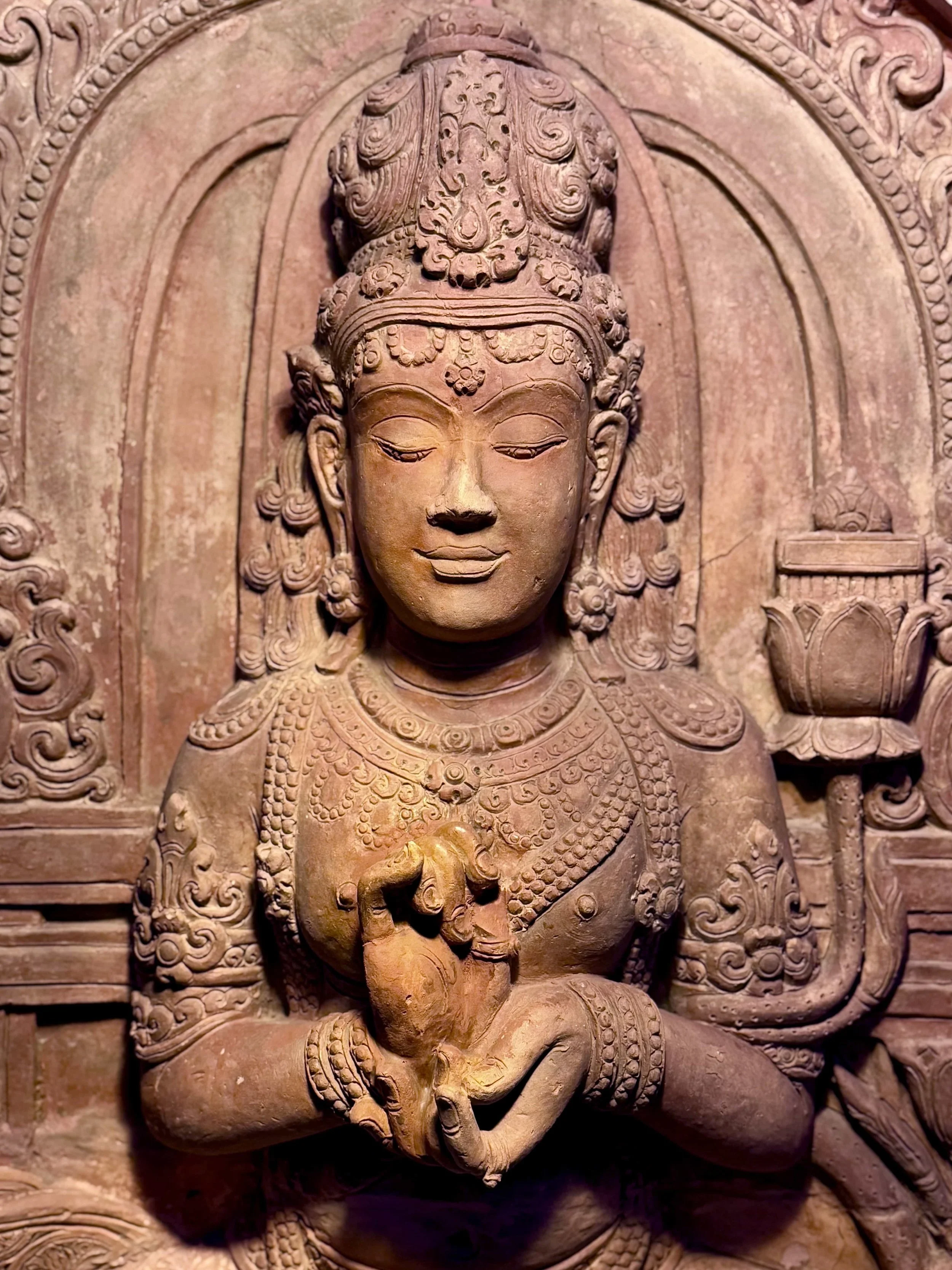 Close-up of a detailed ancient stone carved statue of a deity or figure with ornate jewelry and a serene facial expression, from a historical temple or monument.