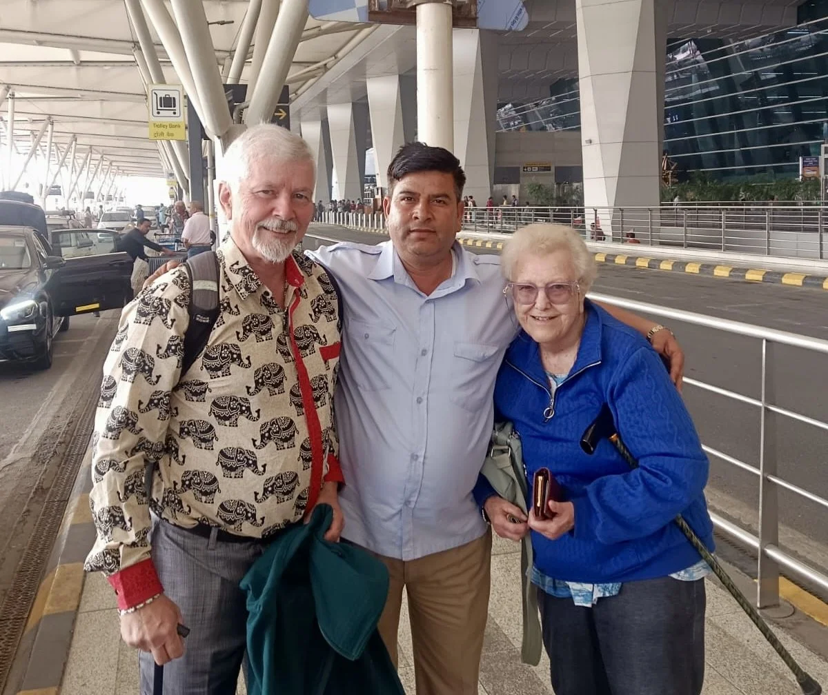 Three people, two men and one elderly woman, standing close together at an airport terminal with cars and people in the background.