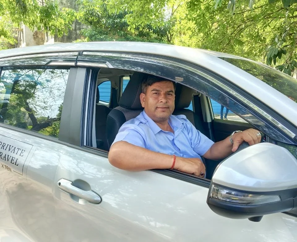 A man sitting inside a white car, wearing a light blue shirt, and looking toward the camera with trees and greenery in the background.