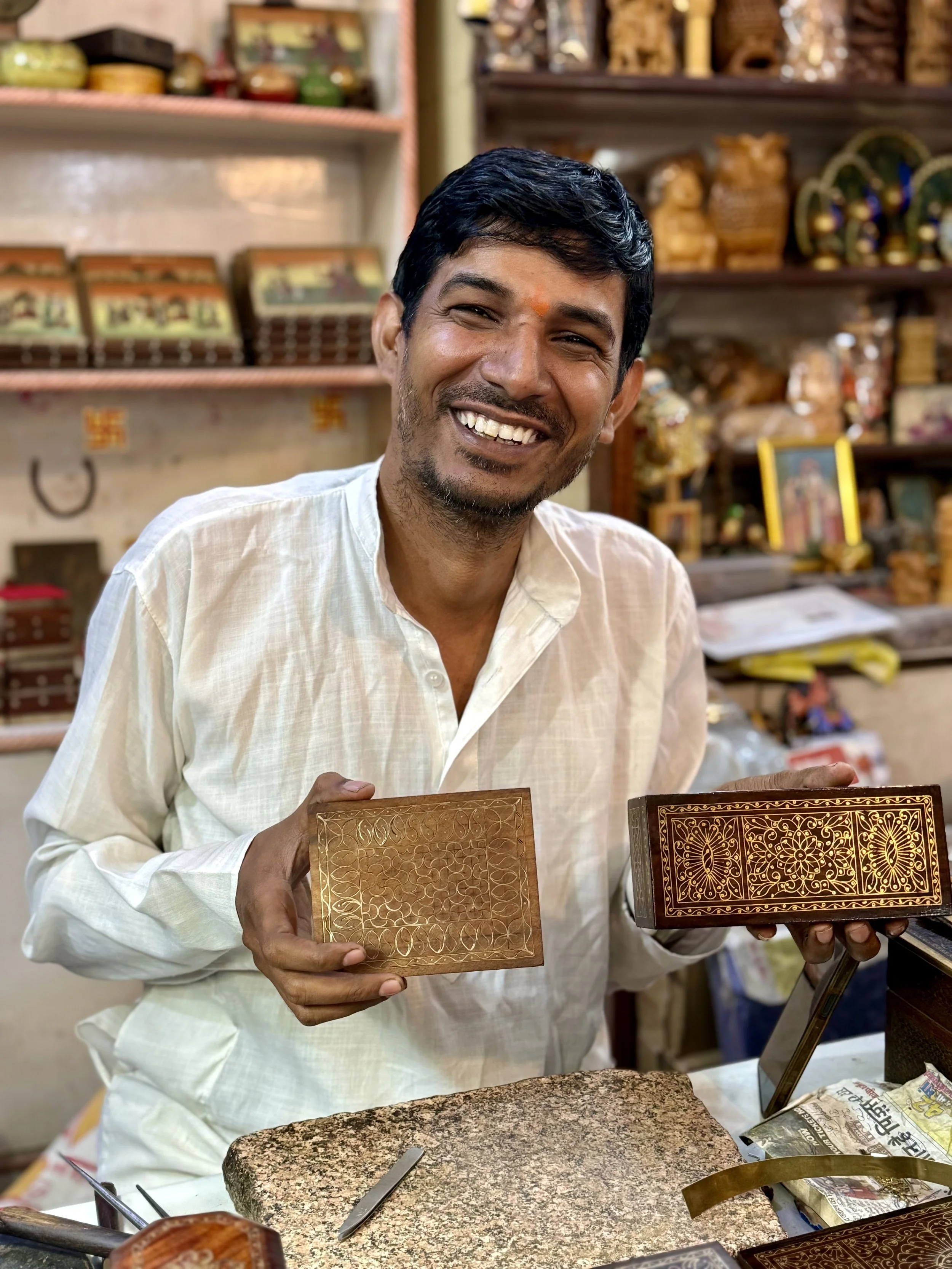 Smiling man in traditional Indian attire showing decorative wooden boxes inside a craft shop.