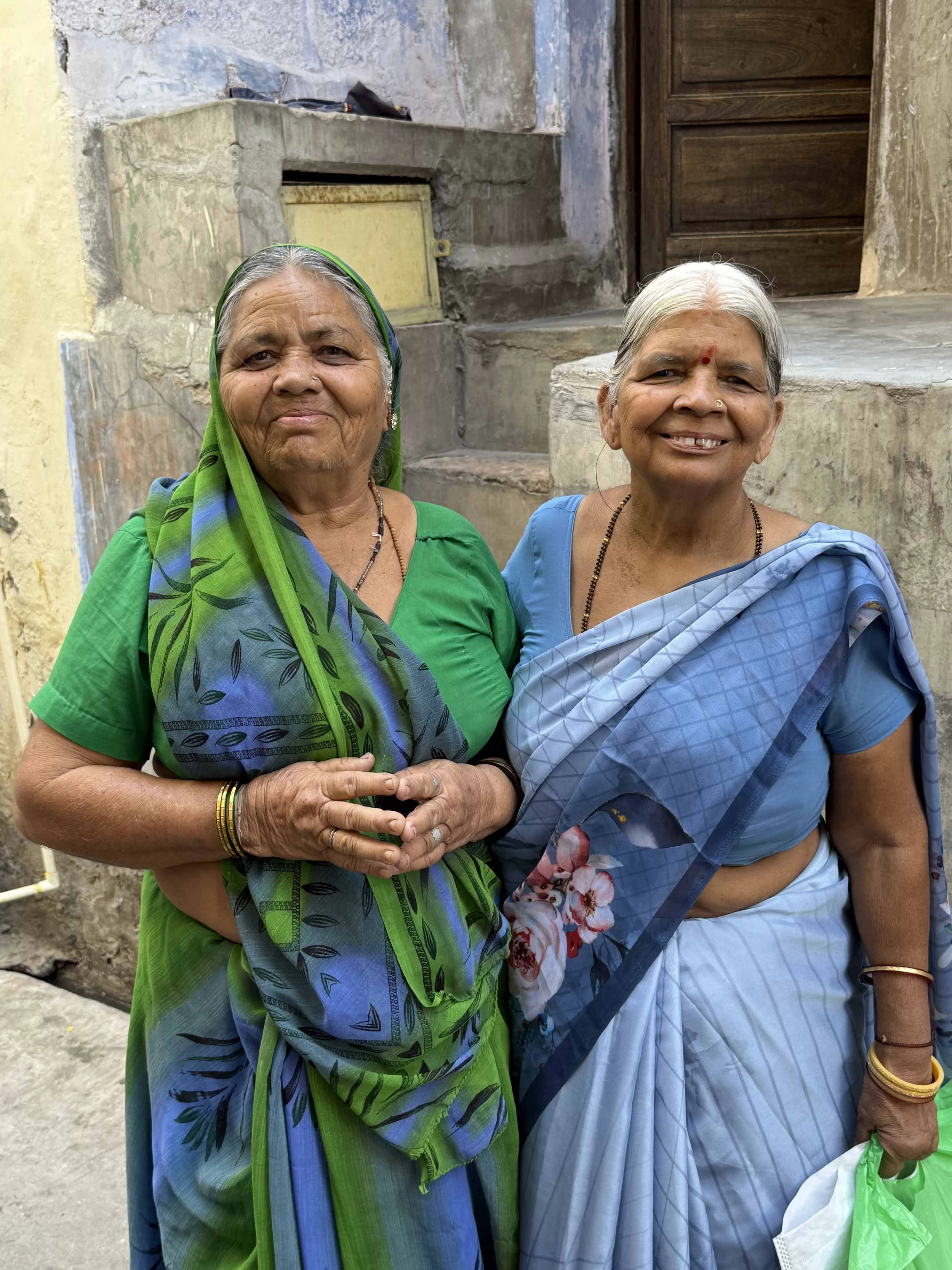 Two elderly Indian women wearing traditional sarees, smiling, standing outdoors in front of a wall and a wooden door.