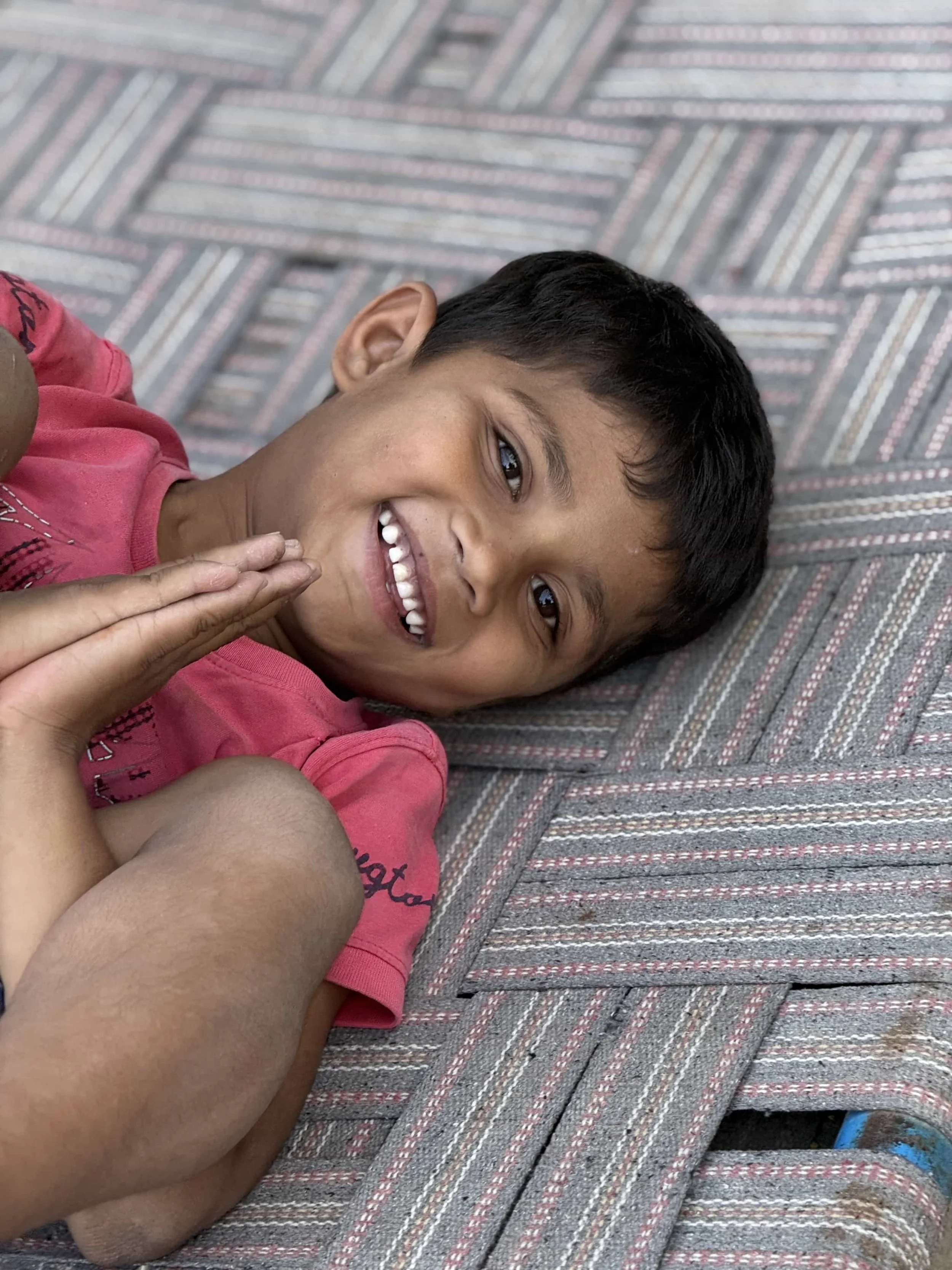 A young boy lying on a striped mat, smiling and looking at the camera. He has short black hair and is wearing a pink shirt.