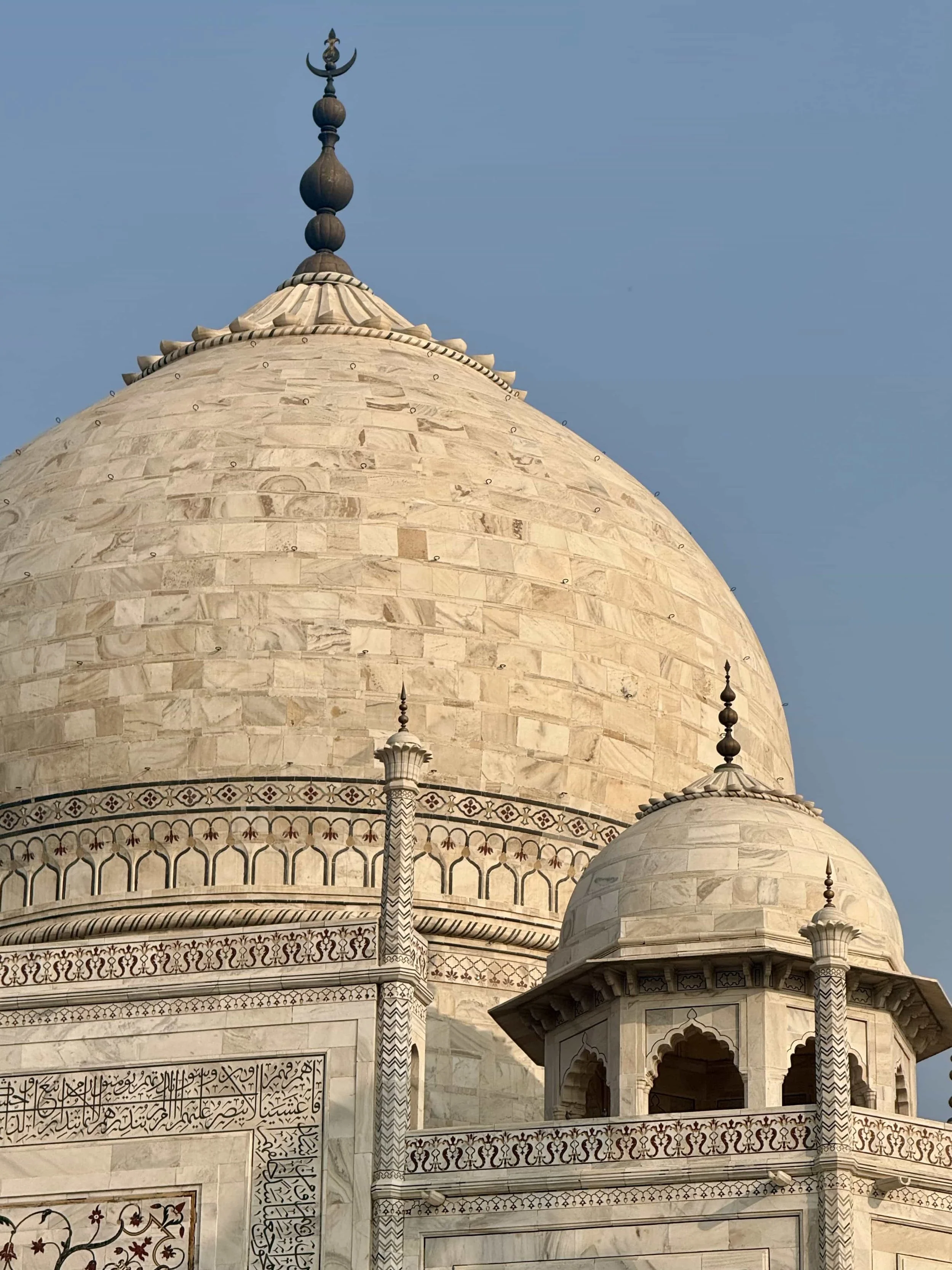 Close-up of the white marble dome of the Taj Mahal with intricate black and white border designs, decorative minarets, and a blue sky in the background.