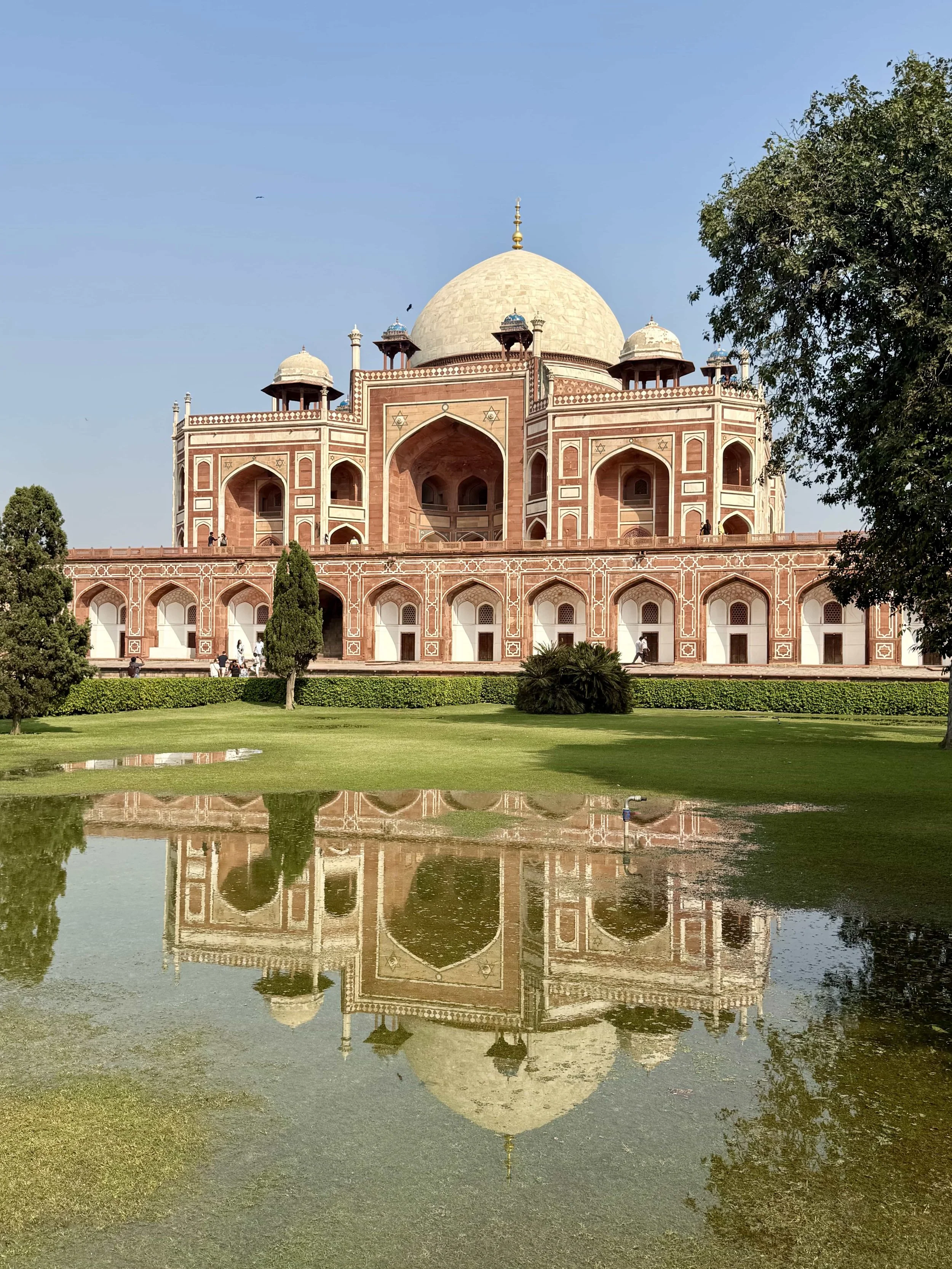 The image shows the Taj Mahal, an iconic white marble mausoleum located in India. It is reflected in a large pond in the foreground, with greenery and trees surrounding the area and a clear blue sky above.