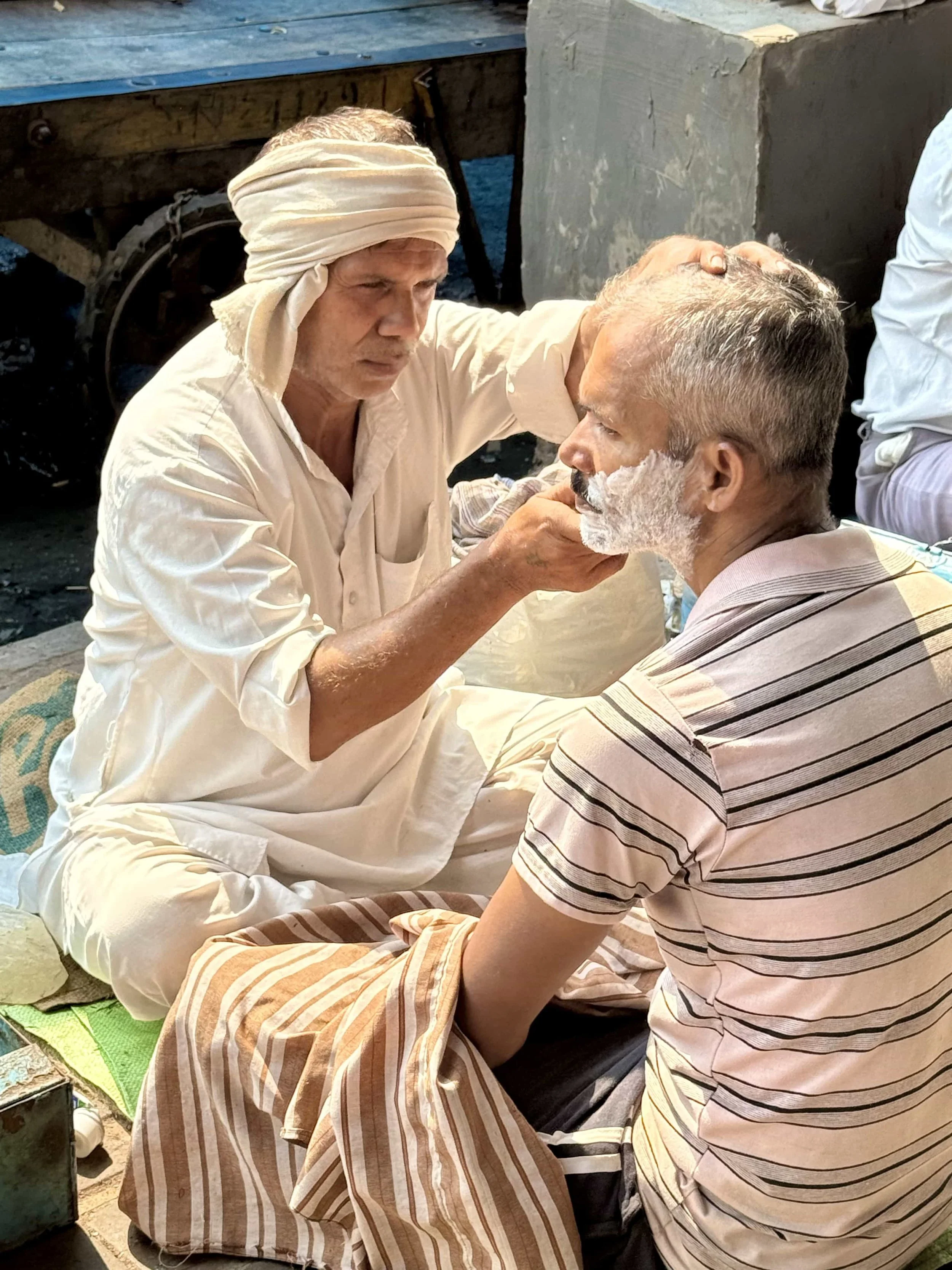 An elderly man getting a shave from another man outdoors, with a wooden cart and concrete structure in the background.