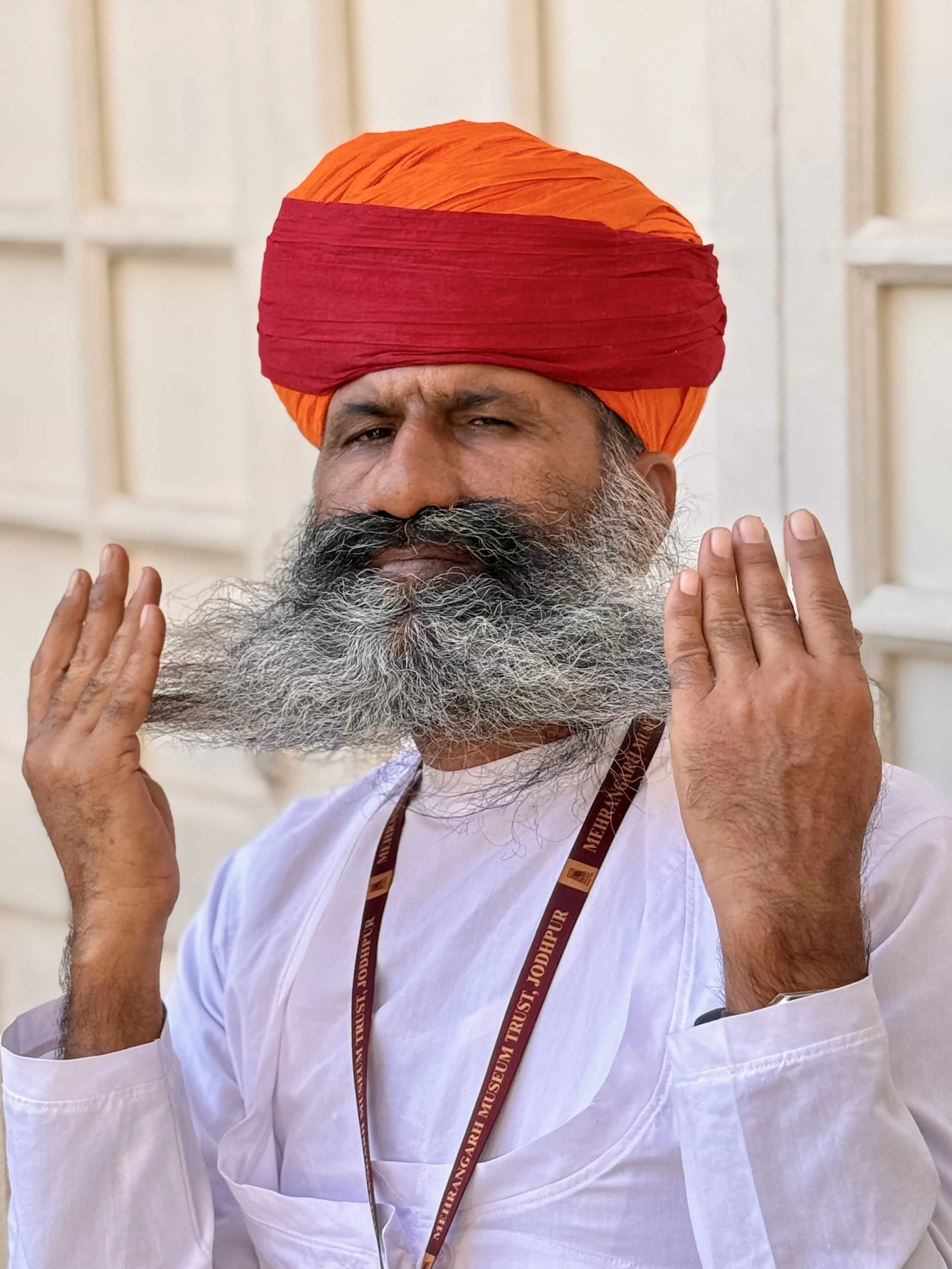 A man wearing a red and orange turban, white shirt, and a lanyard, holding his beard with both hands in front of a light-colored wall.