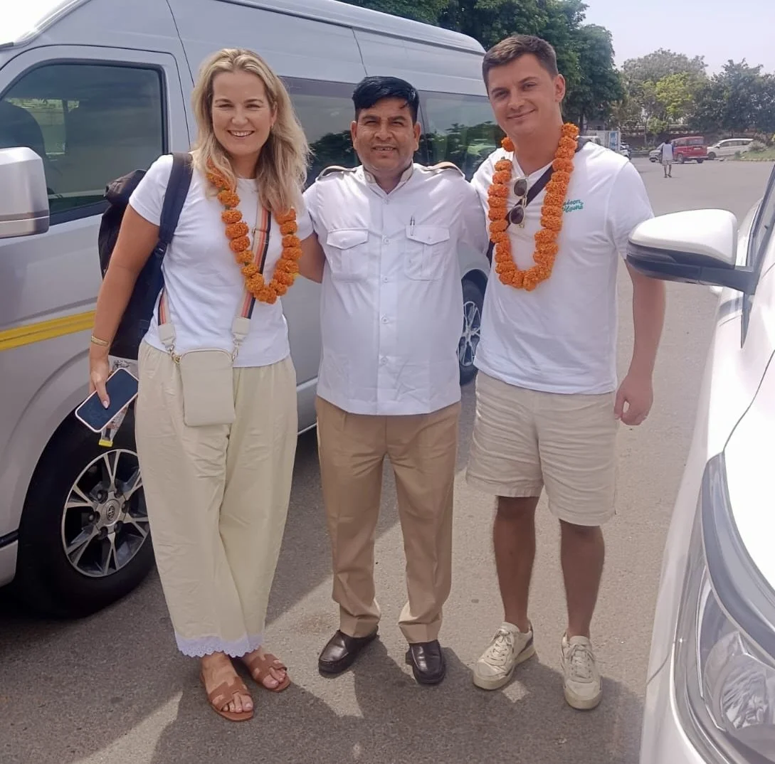 Three people standing outdoors near vehicles, wearing white shirts, with two of them wearing orange flower garlands, smiling at the camera.