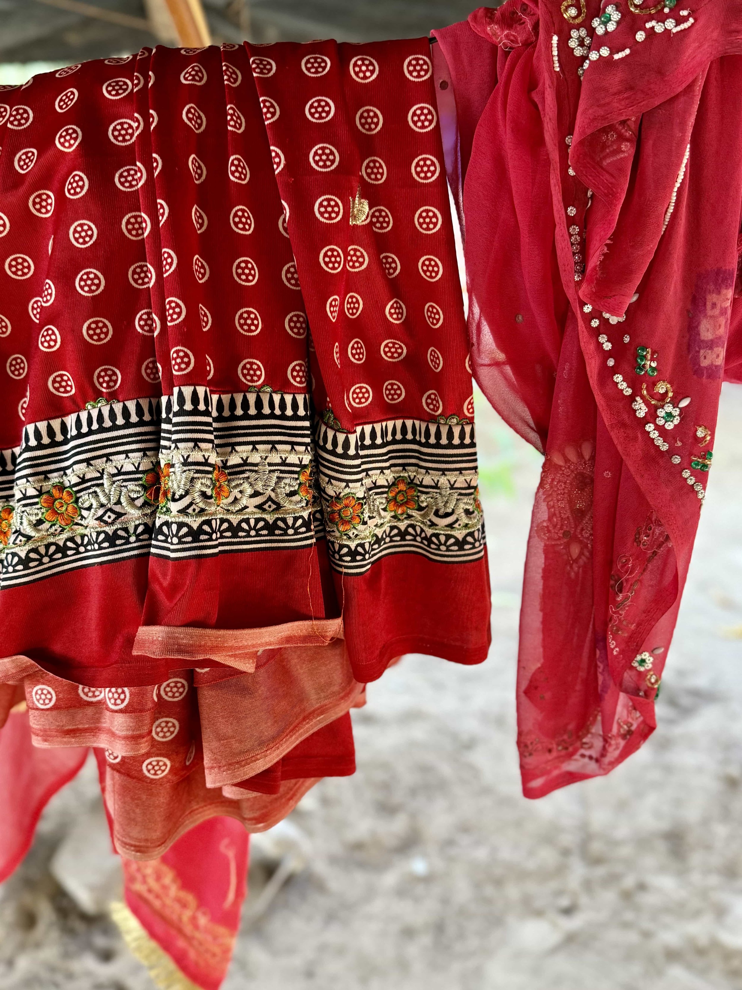 Close-up of hanging red traditional Indian clothes with intricate patterns and embroidery, displayed outdoors with blurry ground in background.