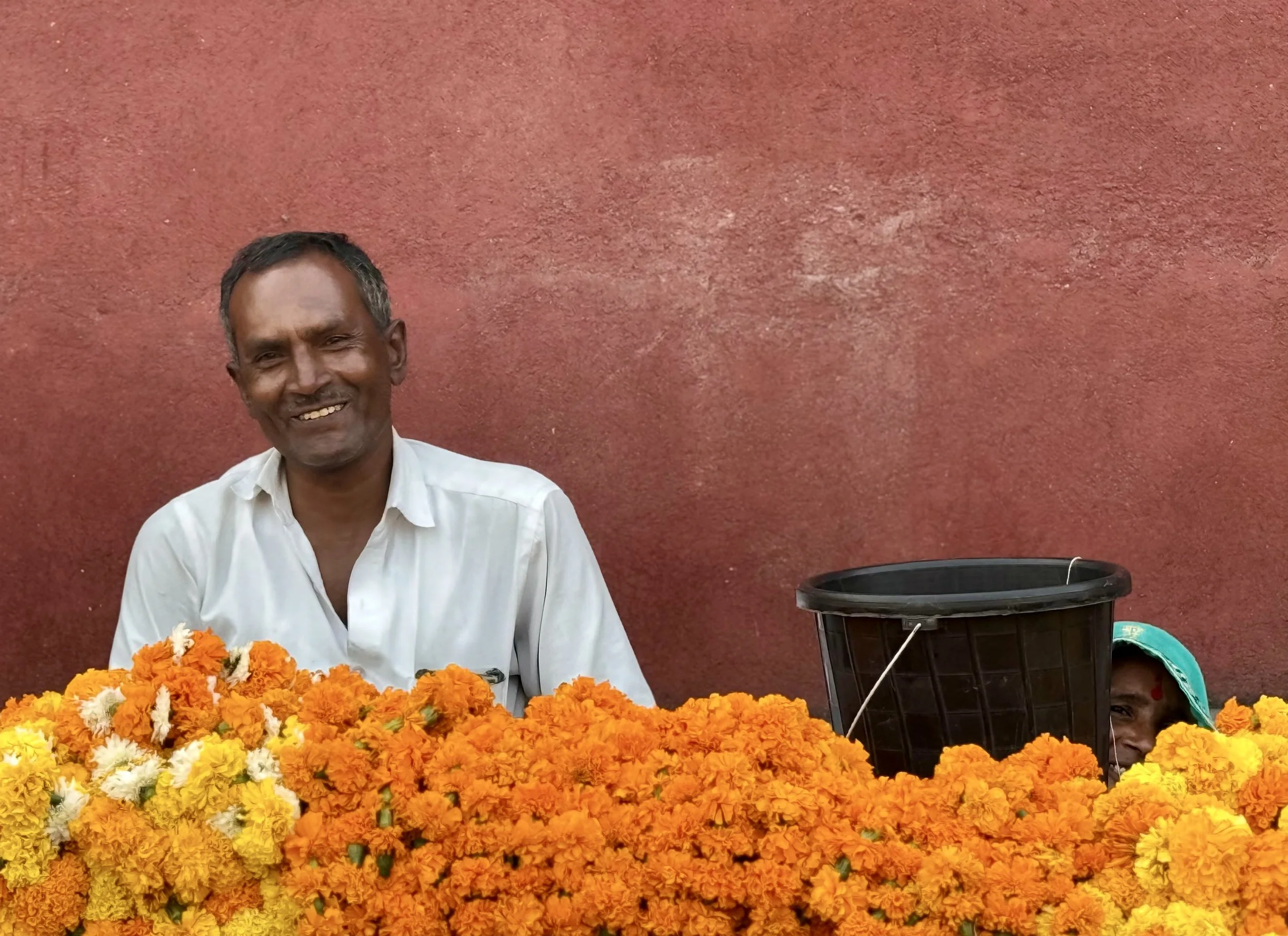 A man smiling in front of a red wall, surrounded by orange marigold flowers, with another person partially visible behind flowers.