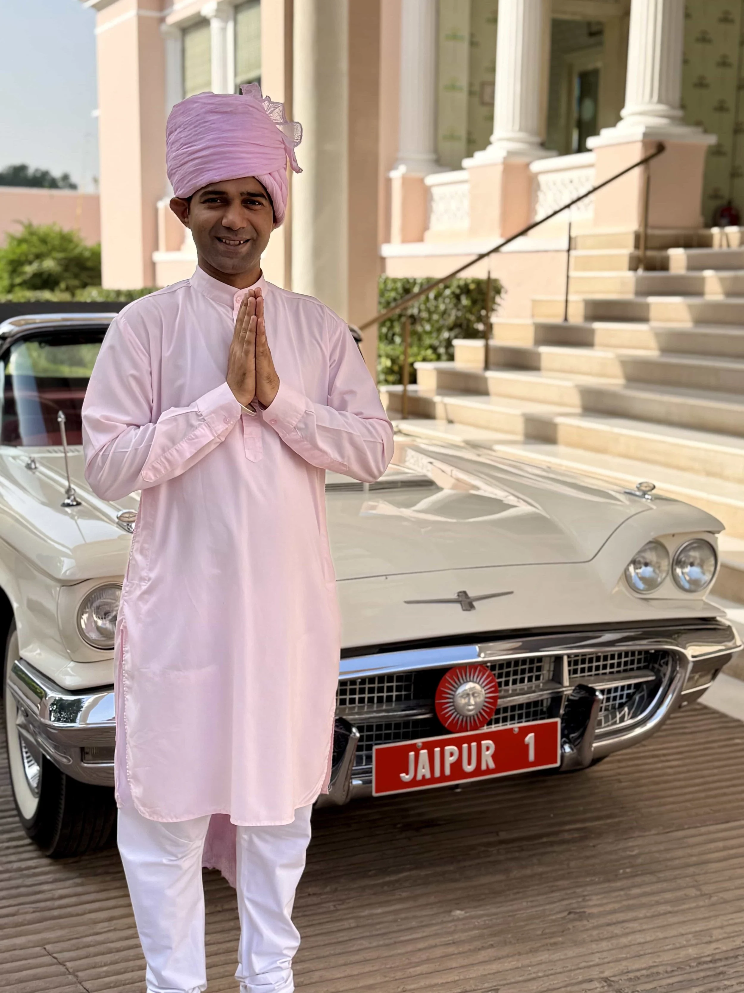 A man dressed in traditional Indian attire with a pink turban and a light pink kurta, standing in front of a vintage white car. The car has a Jaipur license plate and a decorative emblem on the grille. The man is smiling and has his hands in a 'namas