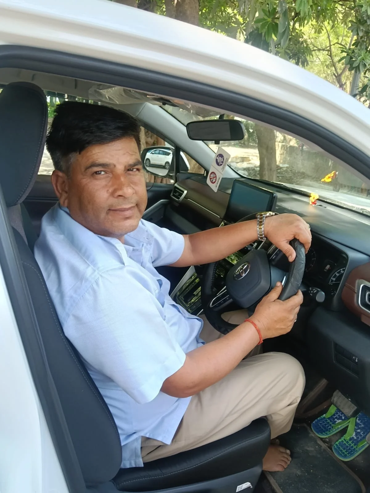 Man sitting in driver's seat of a car, looking at camera, holding steering wheel, wearing a light blue shirt, beige pants, and watch, with trees and another car visible through the window.