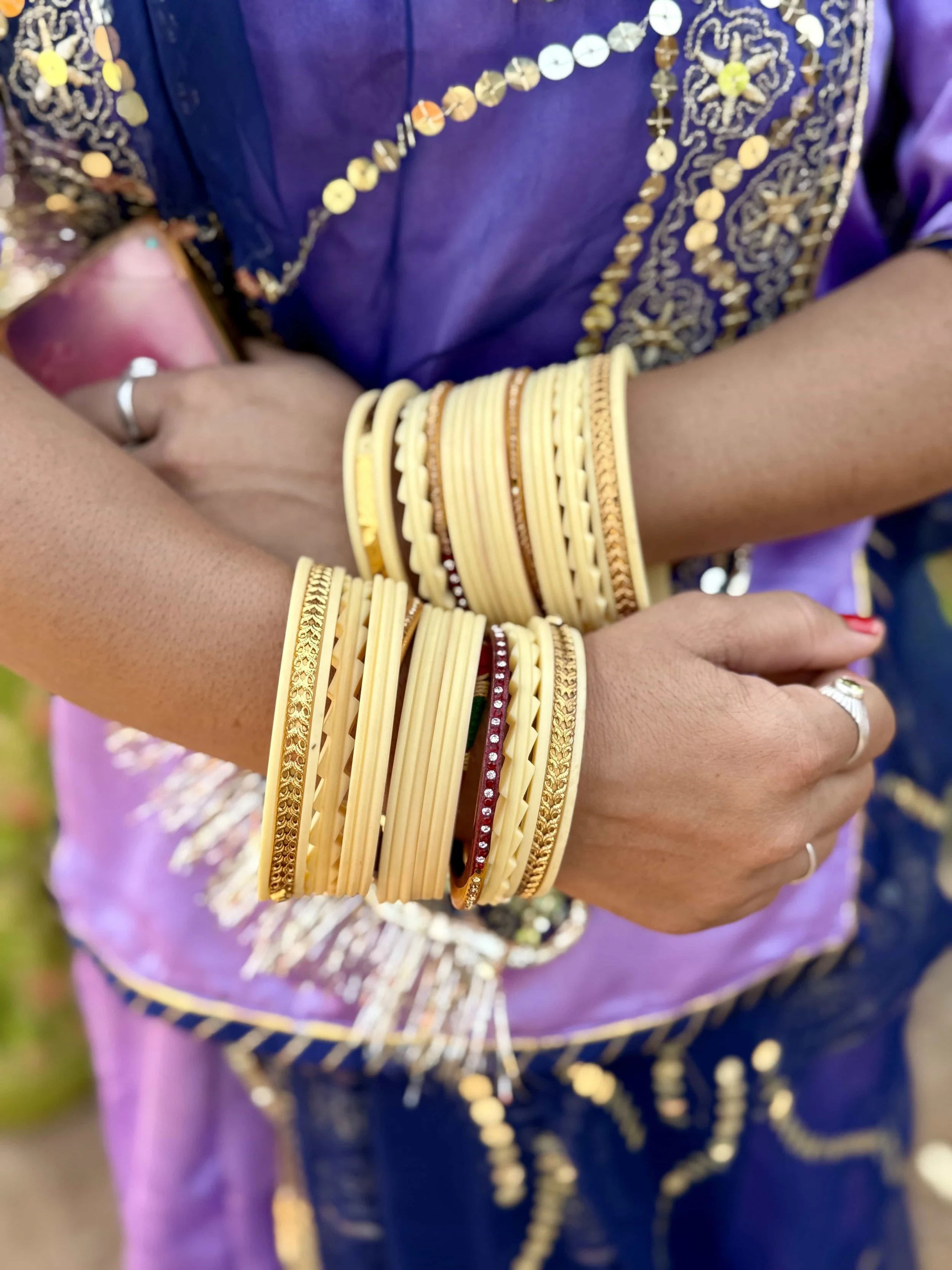 A person wearing traditional Indian attire with purple and gold embroidery, displaying a collection of gold bangles and bracelets on their arm.