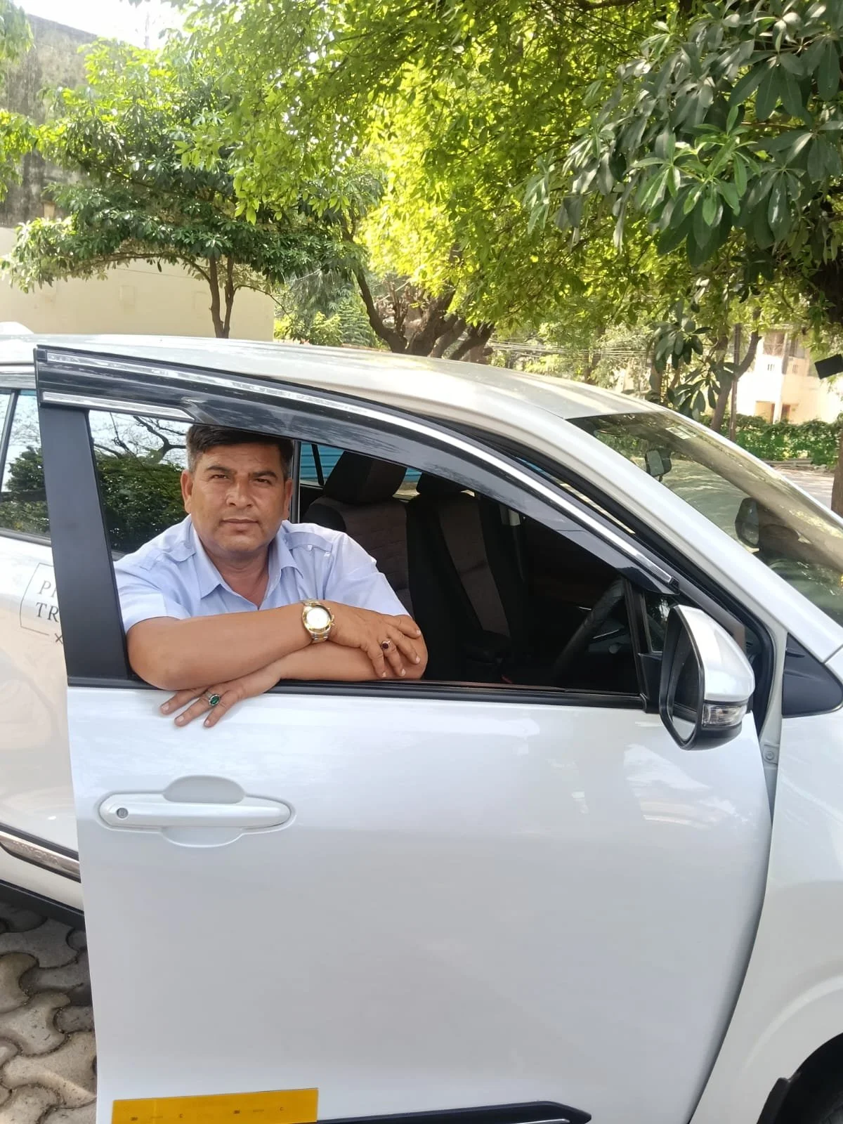 A man with short dark hair wearing a light blue shirt leaning on the open rear door of a white car, with his arms crossed and a watch on his wrist. The background shows green trees and a sunny day.