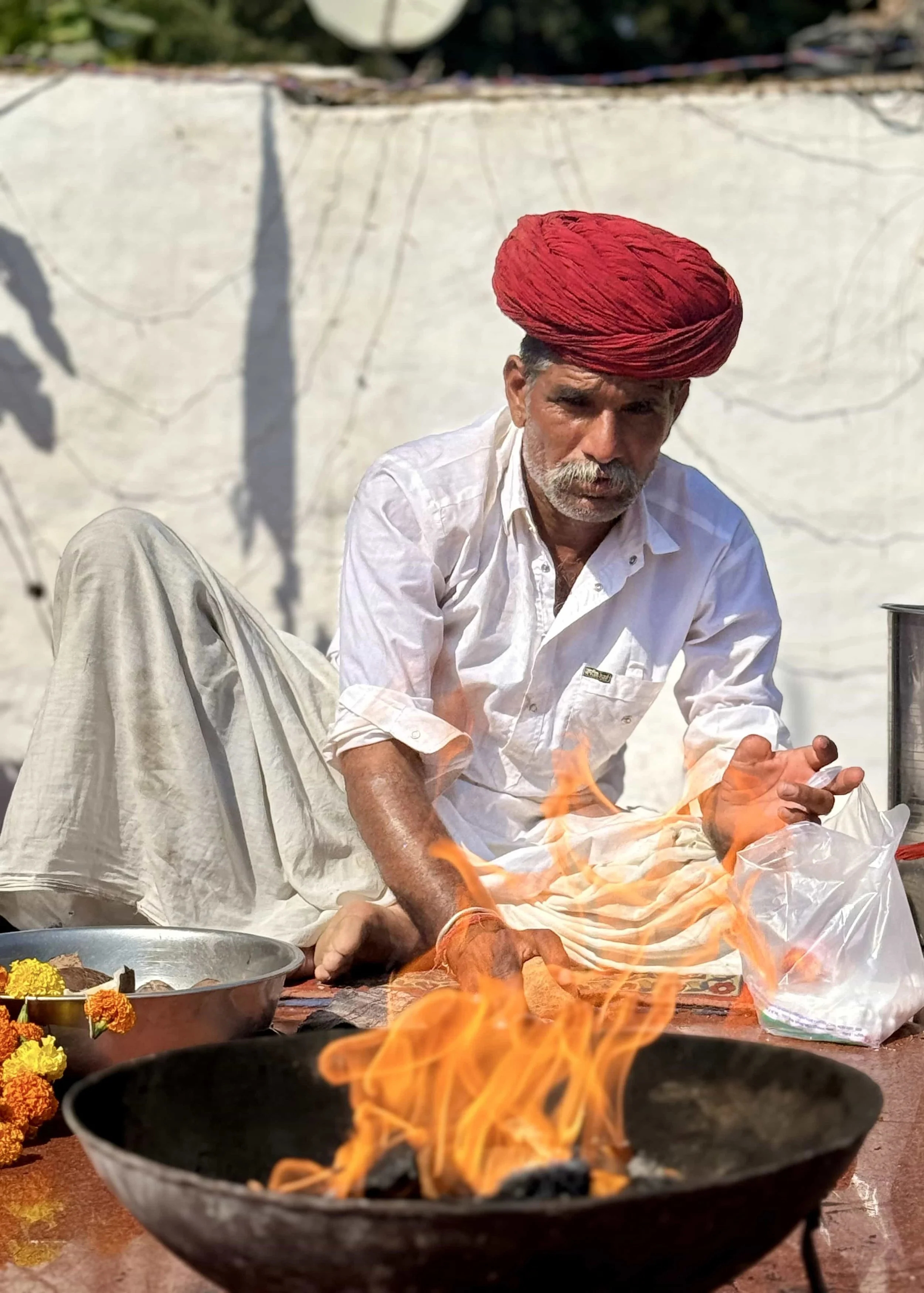 A man wearing a white shirt and a red turban sits on the ground, tending to a fire with flames, surrounded by traditional items and food, against a plain background.