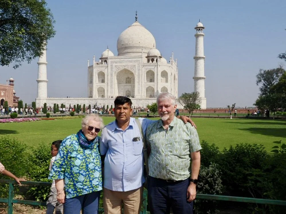 Three tourists standing in front of the Taj Mahal in India, with the iconic white marble mausoleum visible in the background.