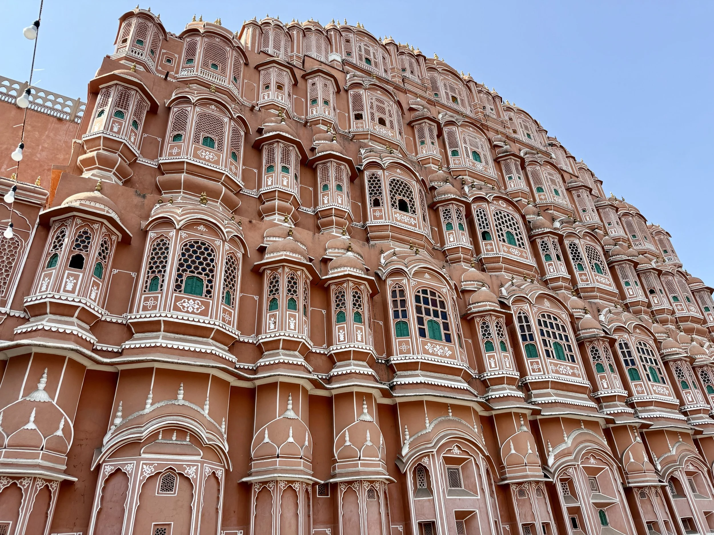 A close-up photo of a large peach-colored building with intricate white and green details, multiple windows with lattice designs and small domed structures, set against a clear blue sky.
