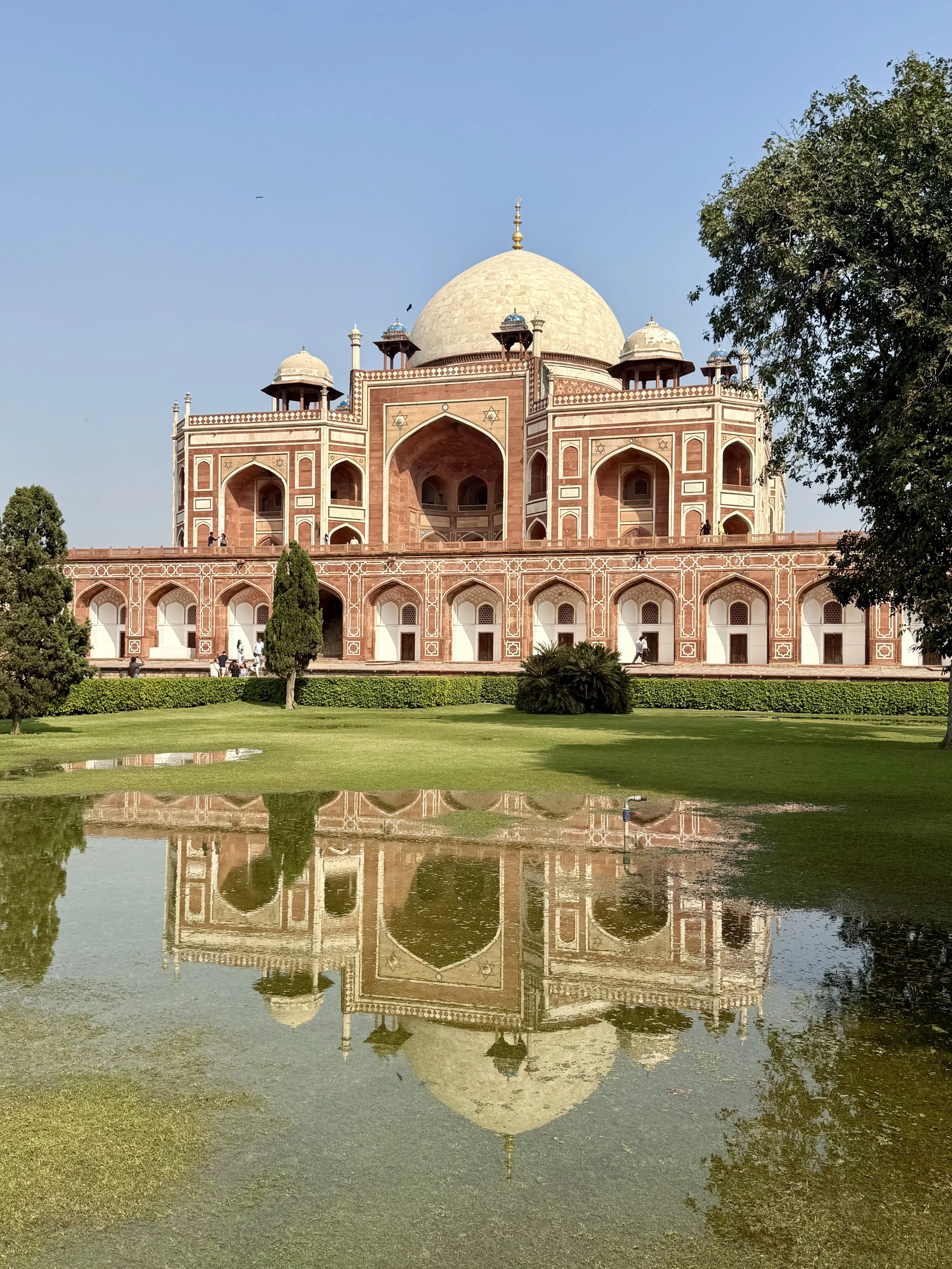 The Taj Mahal, a white marble mausoleum with a large central dome, is reflected in the water in front of it. The sky is clear and blue, with some green trees and grass surrounding the area.
