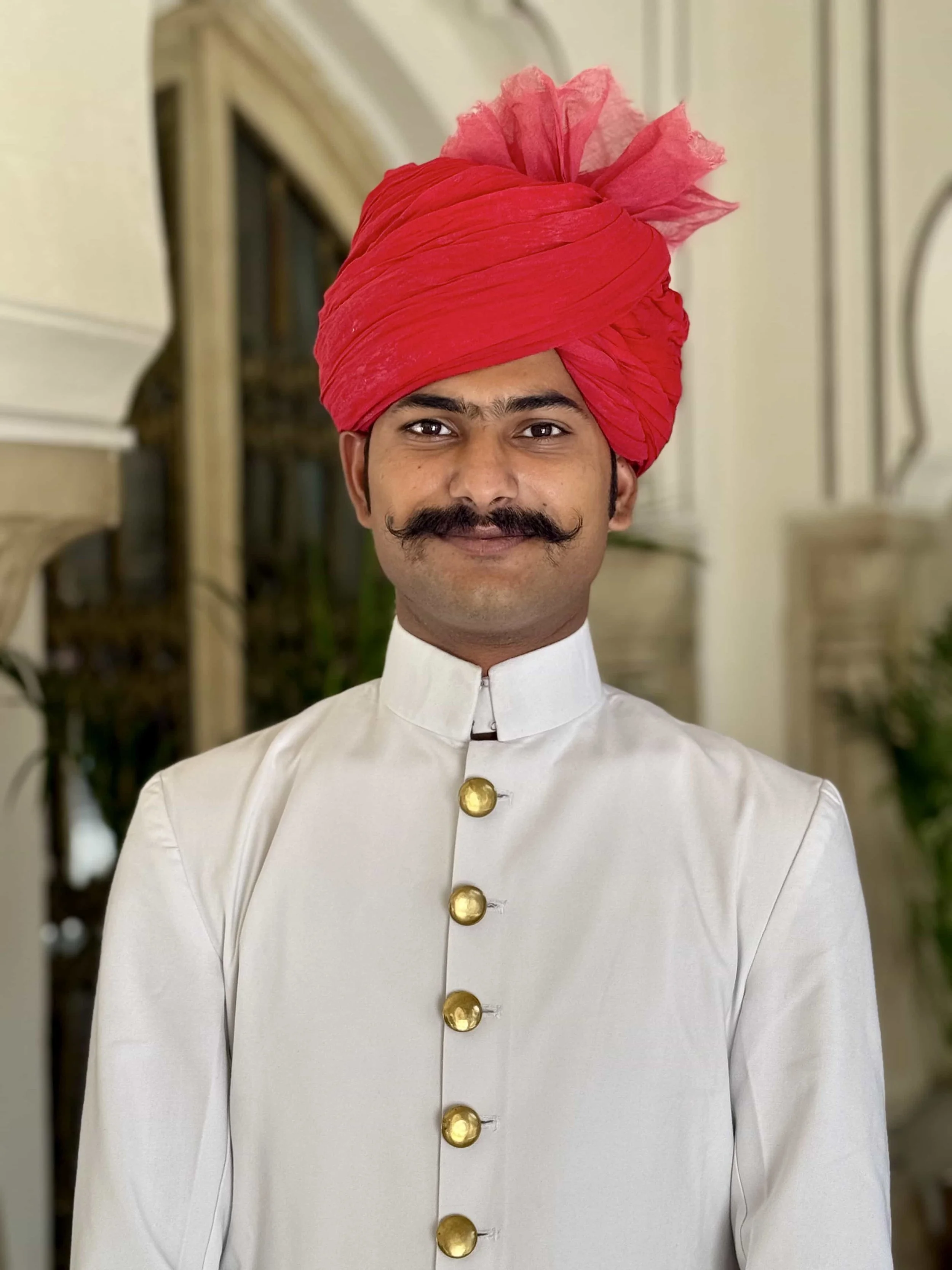 A man wearing a white traditional outfit with gold buttons, a red turban, and a small pink decorative element on top, standing indoors with a blurred background.