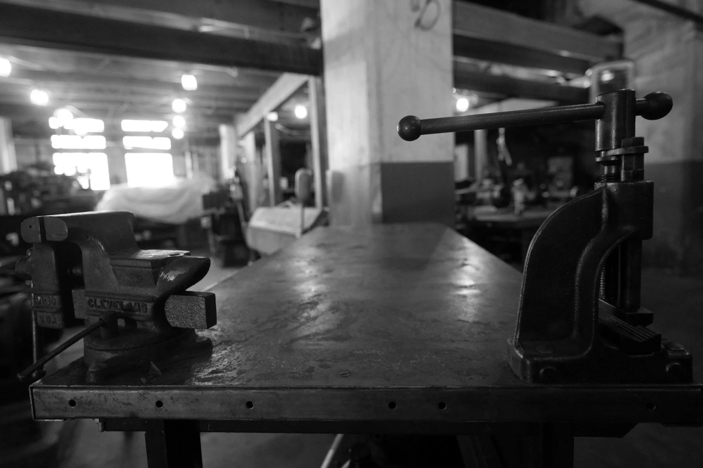 Black and white photo of a workbench in a workshop, with a vise attached to the edge, and various tools and equipment in the background.