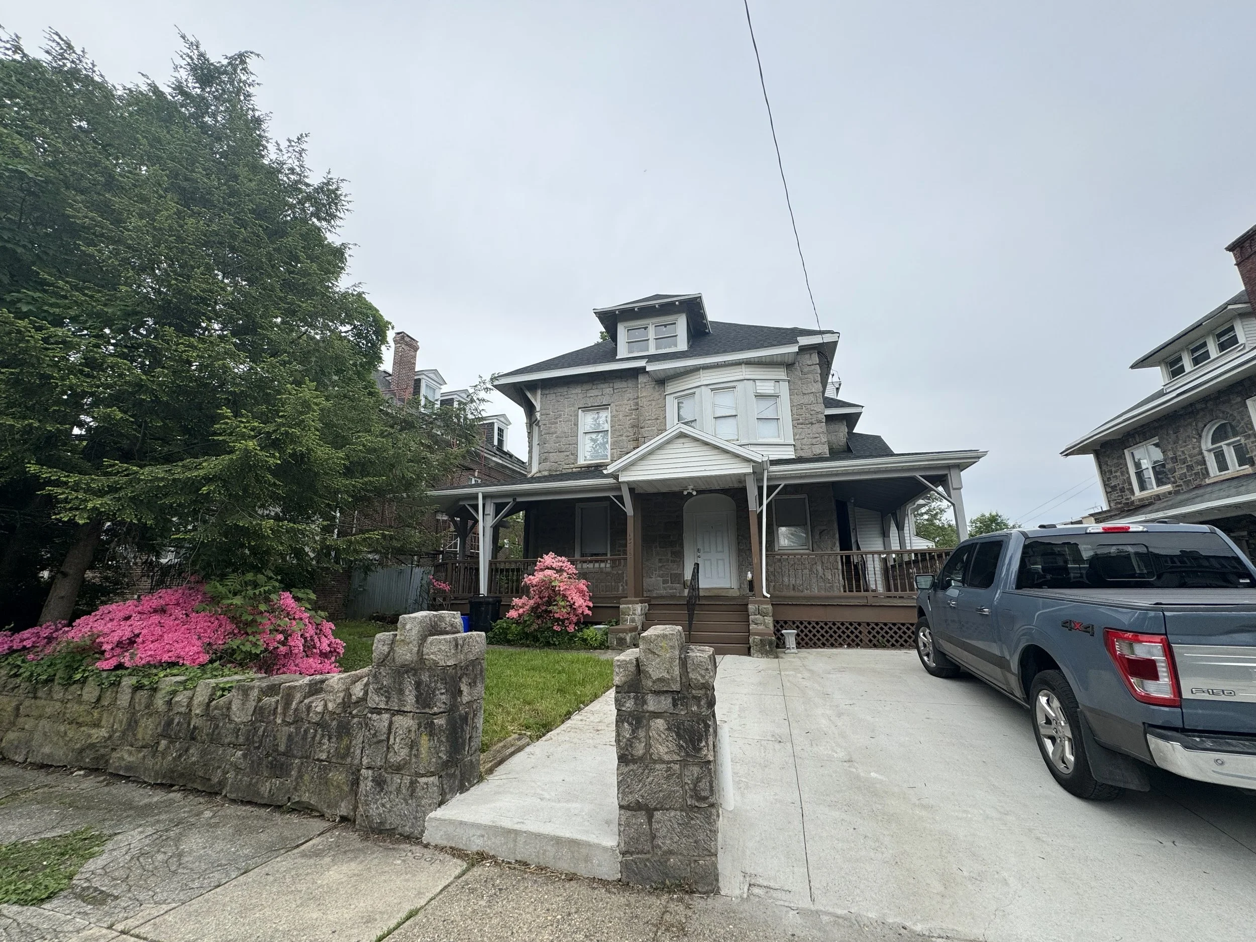 A large, three-story house with a porch and a small set of steps leading to the front door, surrounded by pink flowering bushes and a stone wall, with a driveway and a parked gray truck in front.