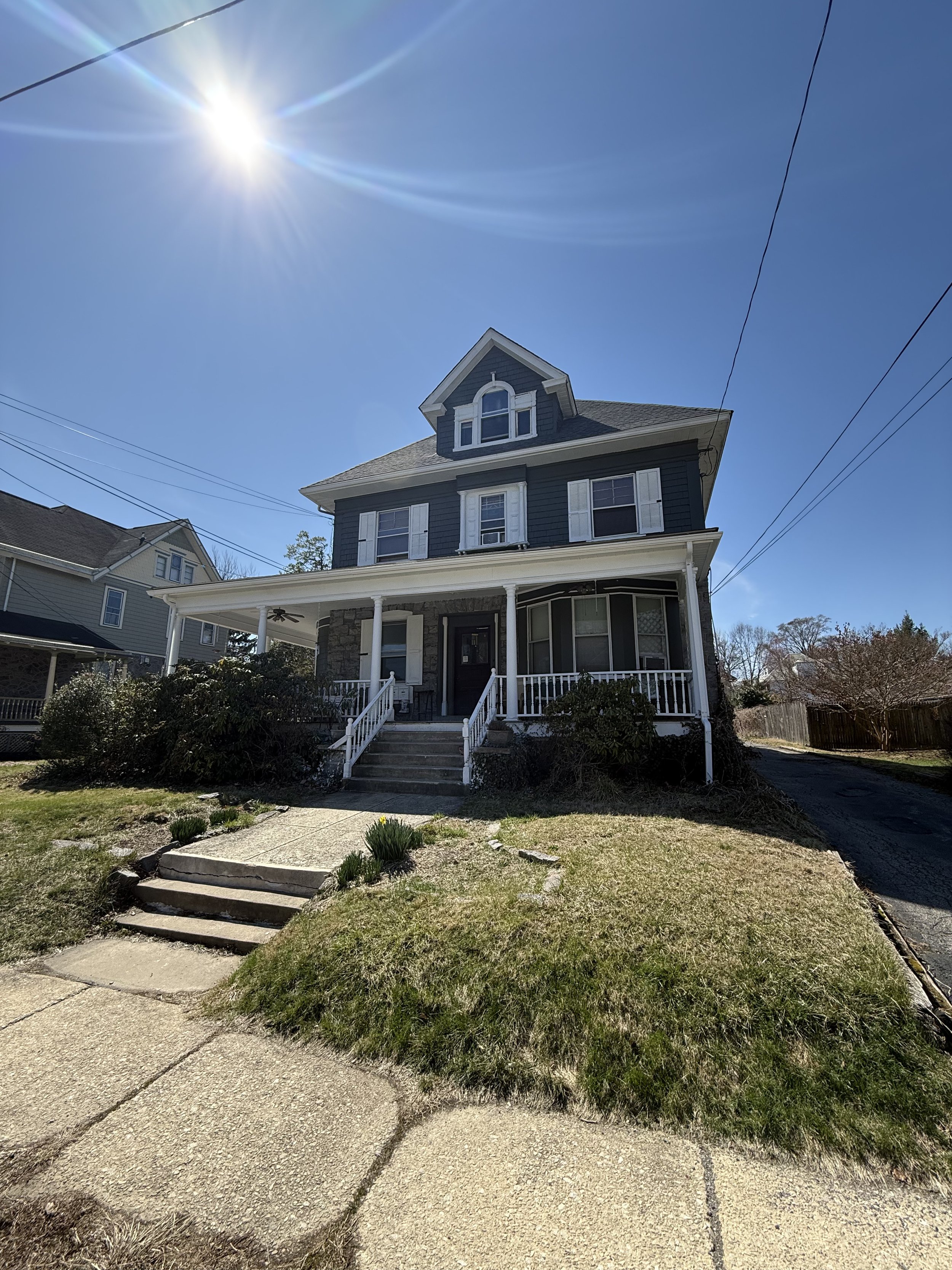 A three-story house with a front porch and stairs, dark blue exterior, and a small dormer window on the top, under a bright sunny sky.