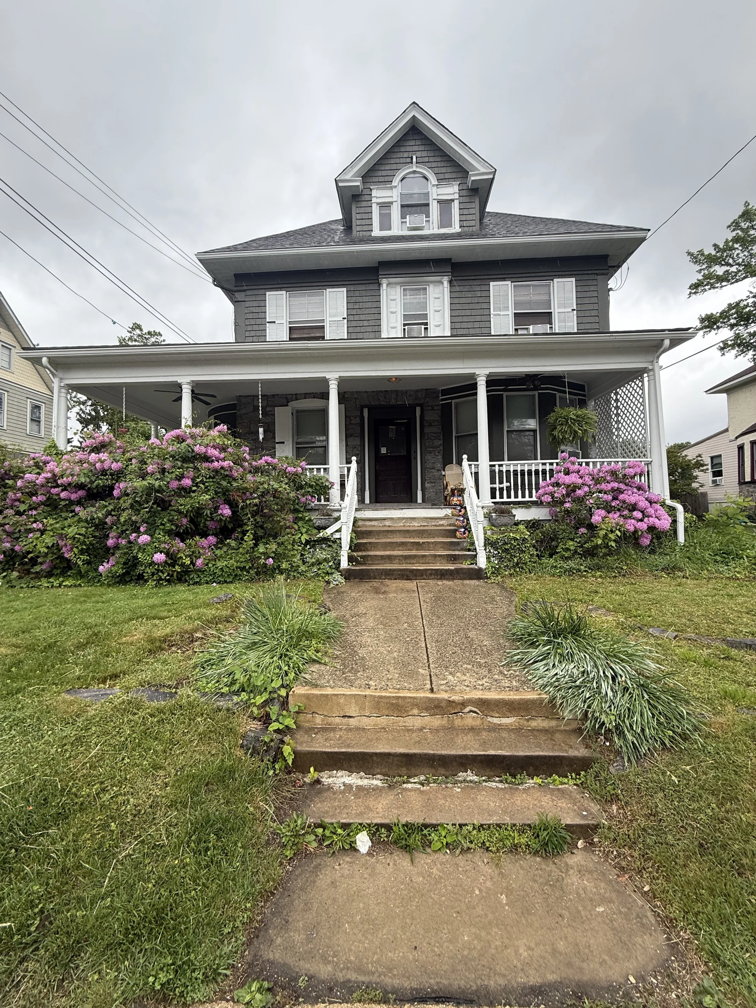 Gray three-story house with a front porch, stairs, lush greenery, and pink flowering bushes.