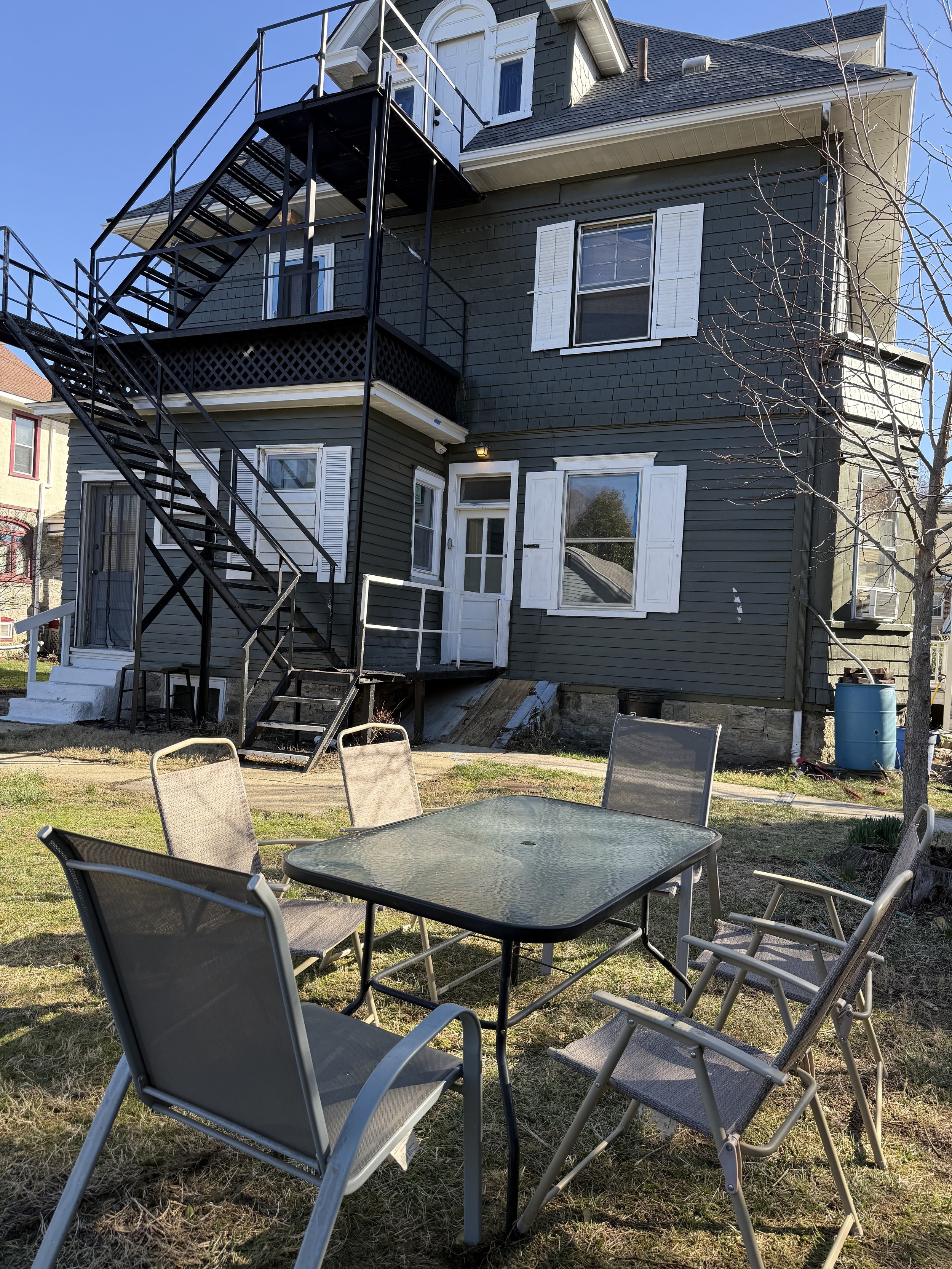 Backyard with a glass patio table and six chairs, adjacent to a dark green three-story house with white window shutters and an external black metal staircase leading to the second floor.