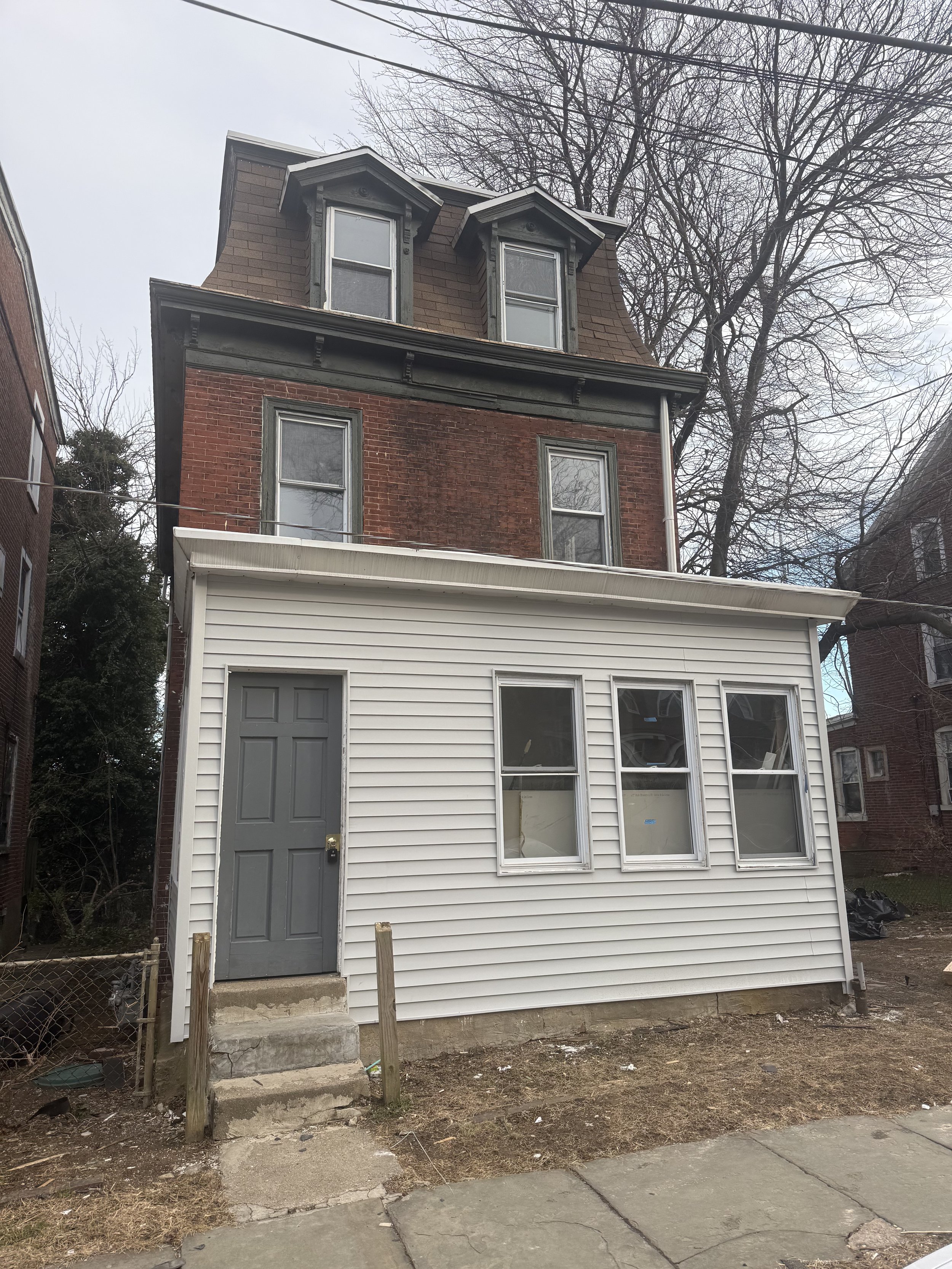 Front view of a multi-story house with white vinyl siding on the first floor, brown brick on the second floor, and dark-colored shingles on the attic level. The house has a gray door, three windows on the first floor, two windows on the second floor,