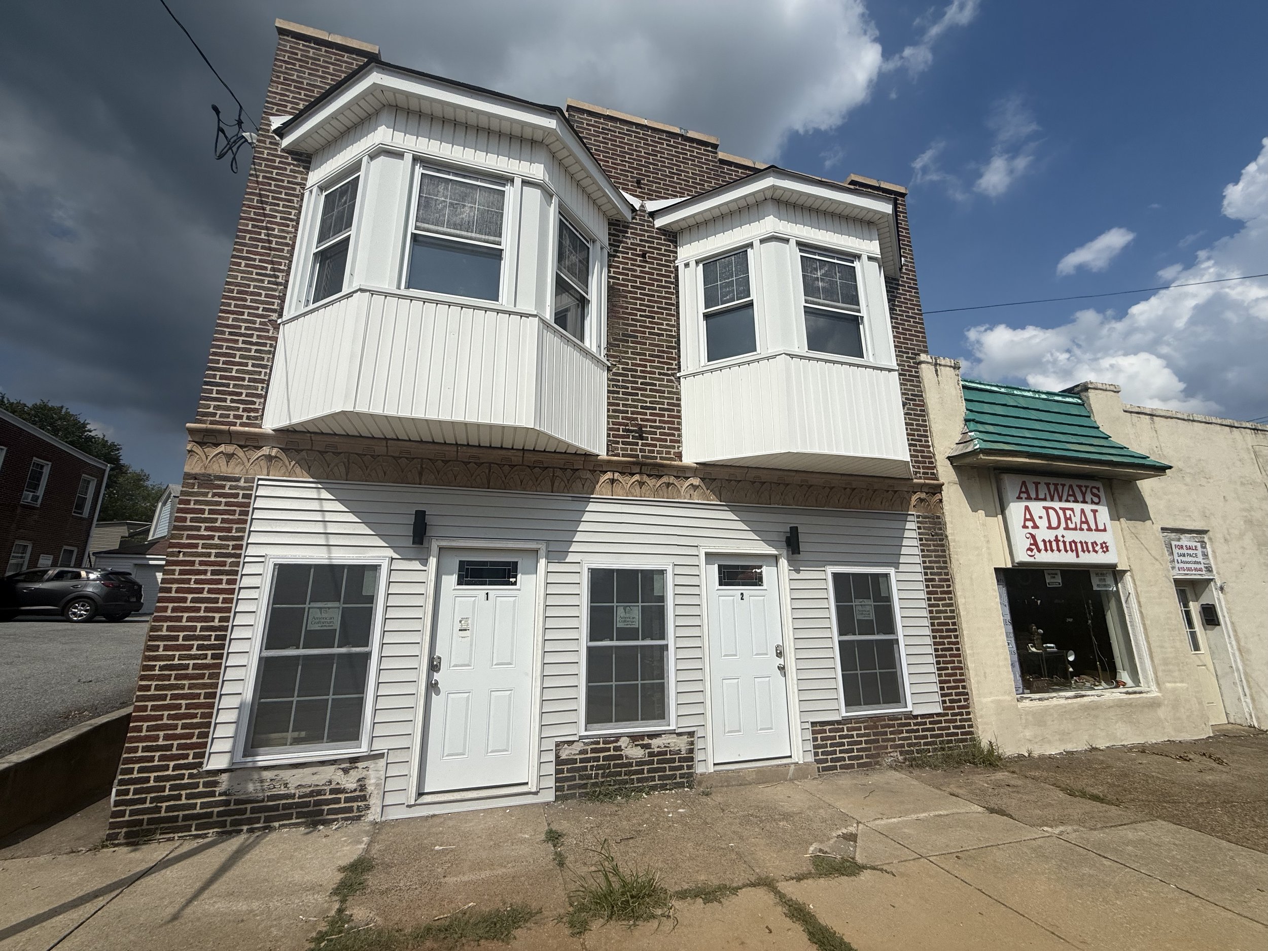 Two-story brick and white siding building with two white doors, windows, and bay windows on the second floor. Adjacent to an antique store with a sign reading 'Always A-Deal Antiques' on a cream wall with a green awning. Overcast sky in the backgroun