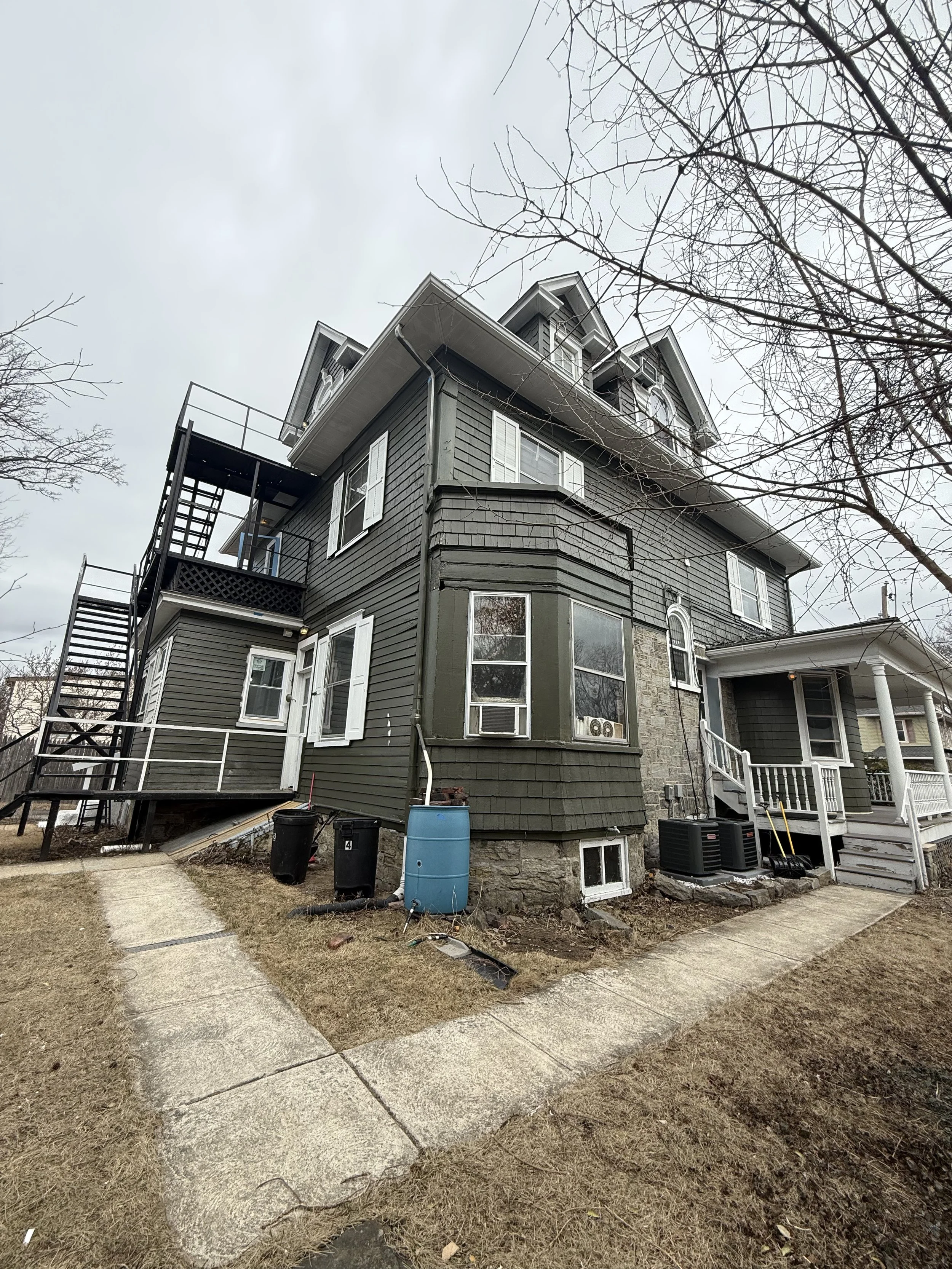 A multi-story, gray-colored house with a front porch, multiple windows, and an external staircase. The yard has a concrete pathway, some bare grass, and several black trash bins near the house.