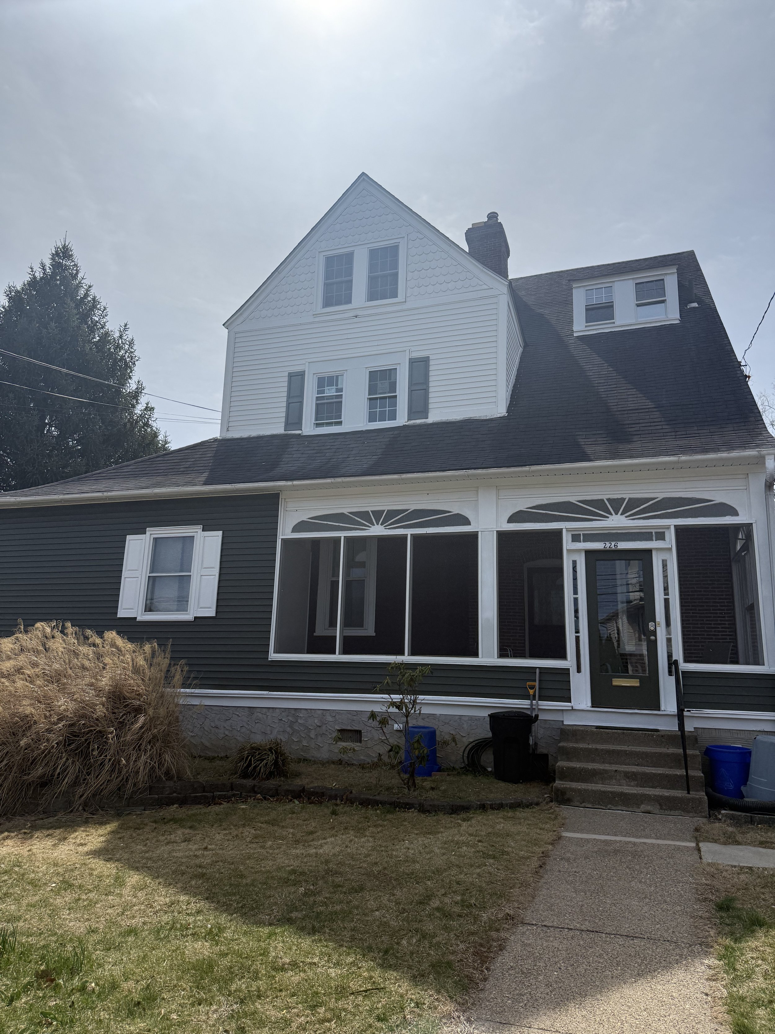 Front view of a house with a porch, steps, small lawn, and various plants, under partly cloudy sky.