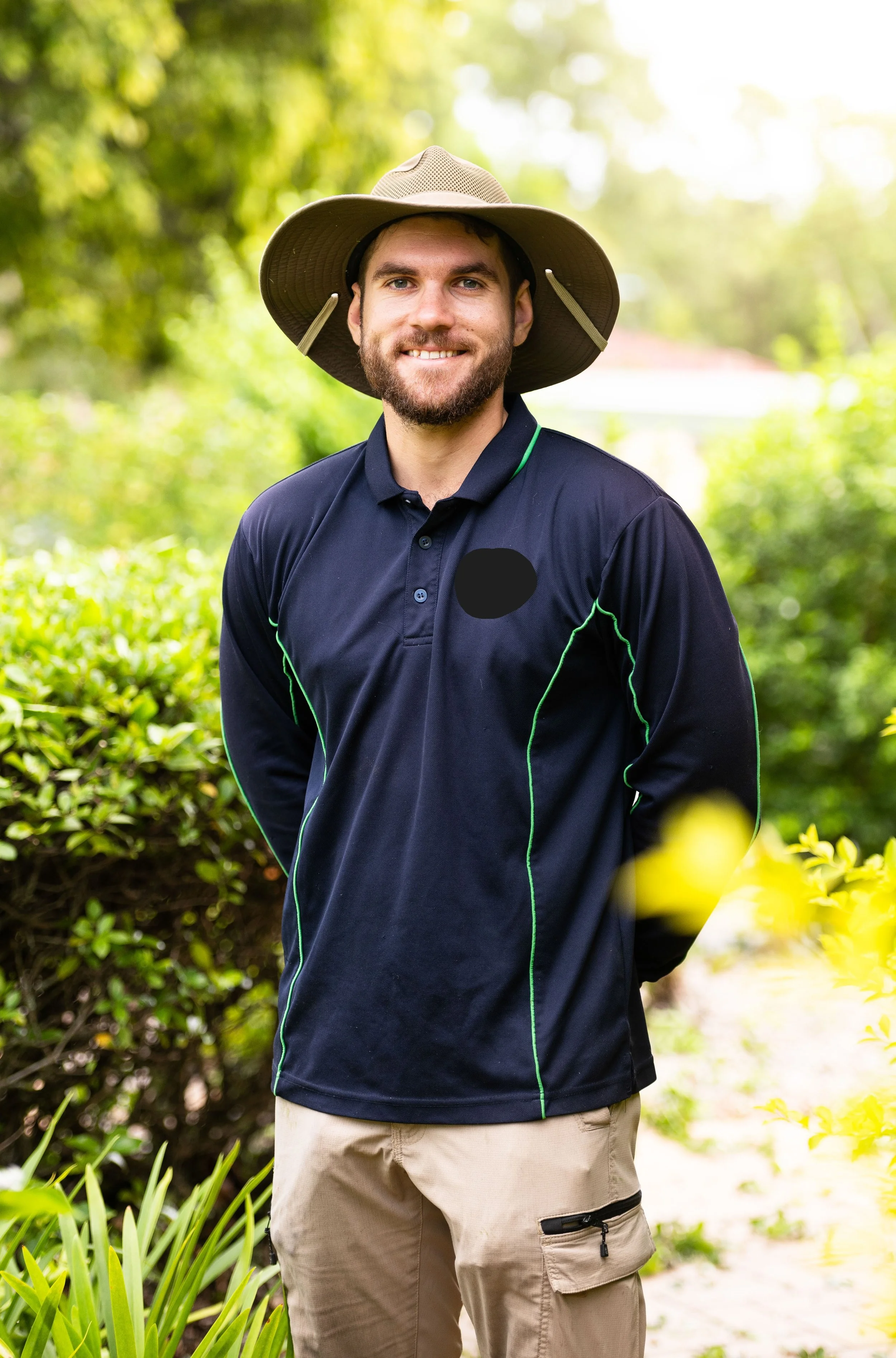 Young man smiling outdoors in a hat, wearing a navy sports shirt with green accents, and khaki cargo shorts, surrounded by lush greenery.