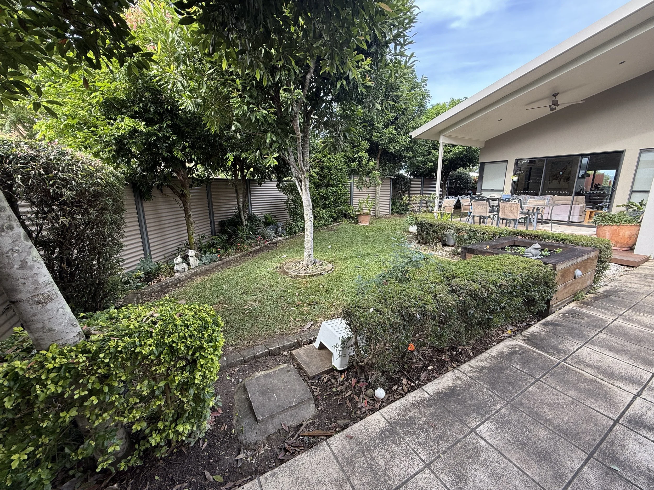 A backyard patio with outdoor dining table and chairs, lawn with trees, shrubs, and garden decorations, and a house with large glass doors.