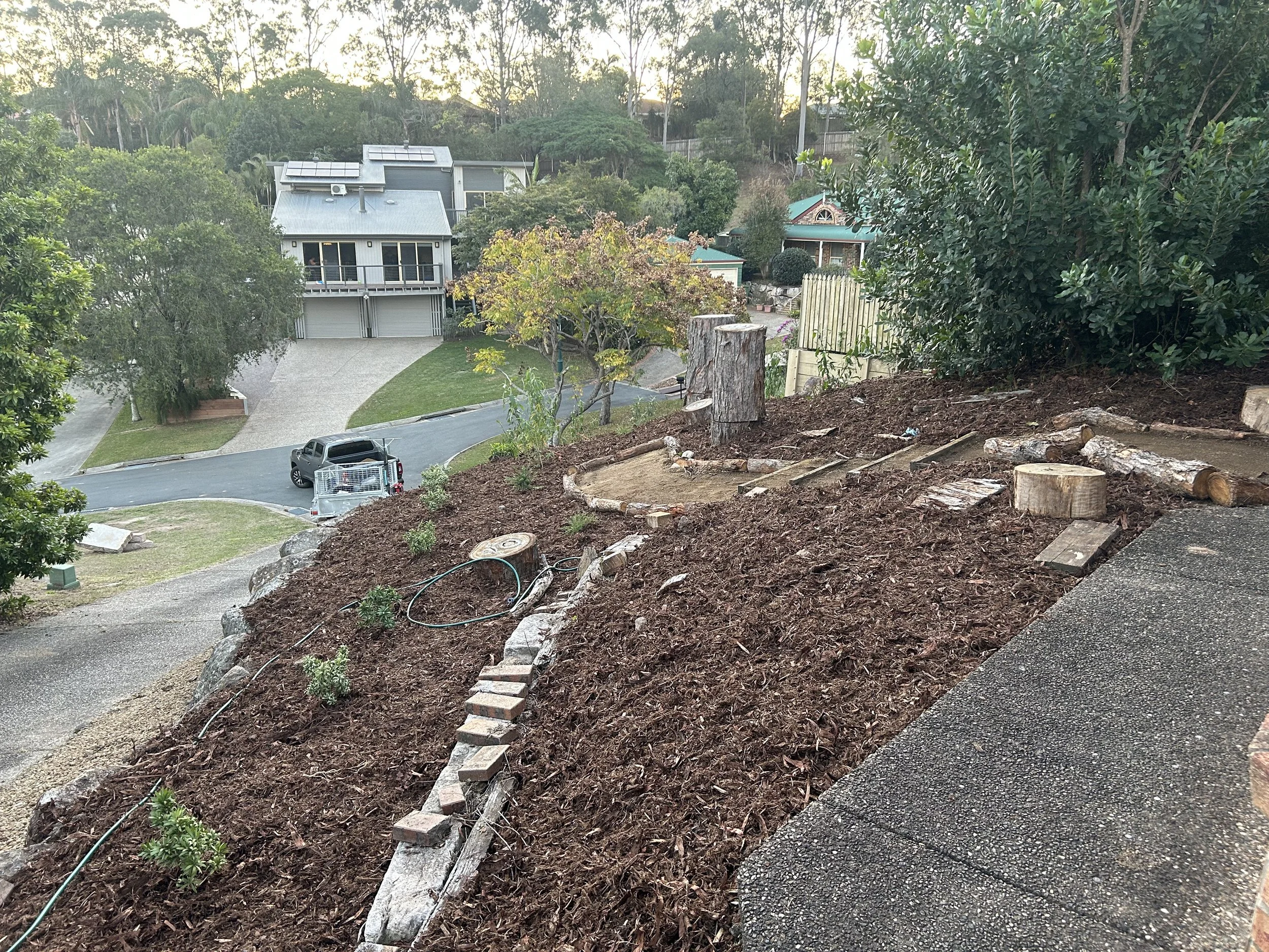 A hillside with freshly turned soil, small plants, and garden bed edging made of rocks and bricks. Logs and a coiled garden hose are also present, with a house and trees in the background.