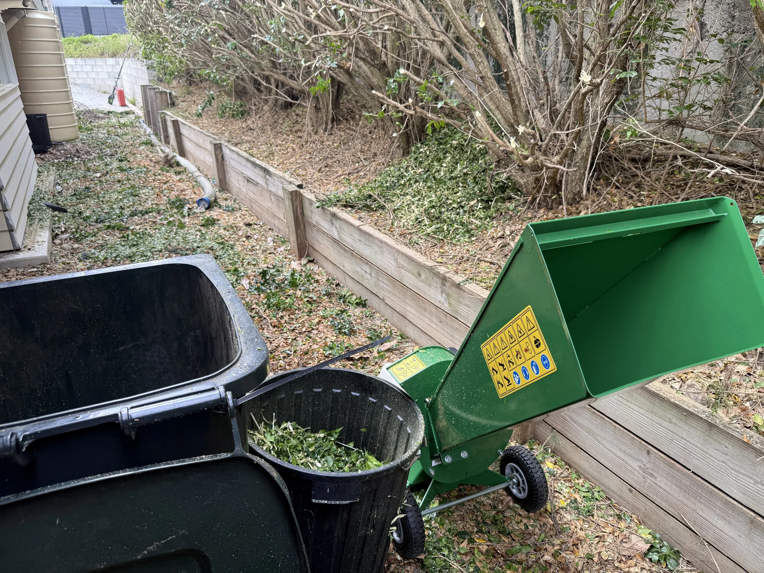 A green wood chipper with a warning label on the side, feeding shredded branches into a black trash bin in a backyard next to a wooden fence and a house.