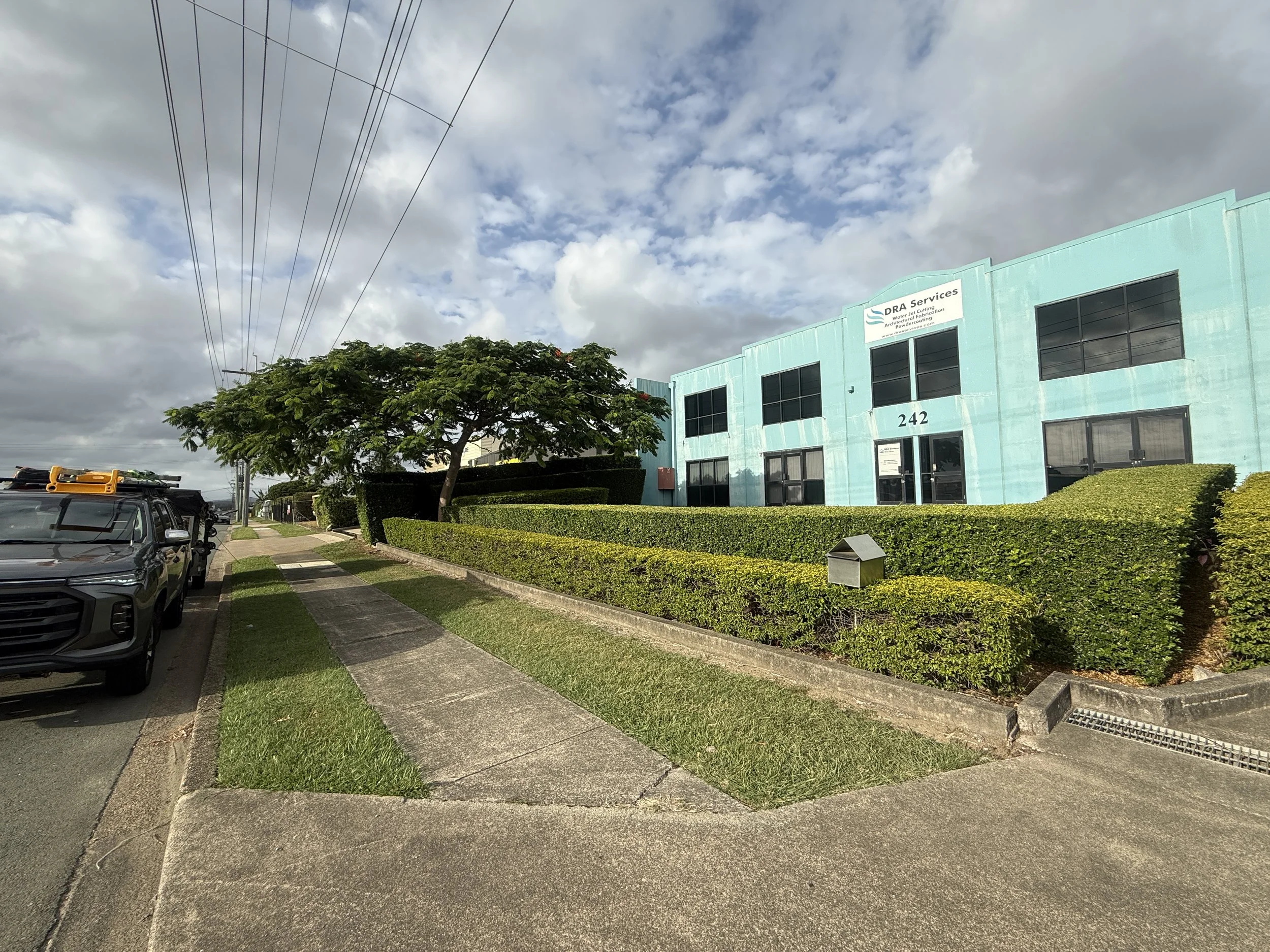 Street view of a light blue commercial building with the number 242, green bushes, a sidewalk, parked cars, and a cloudy sky.
