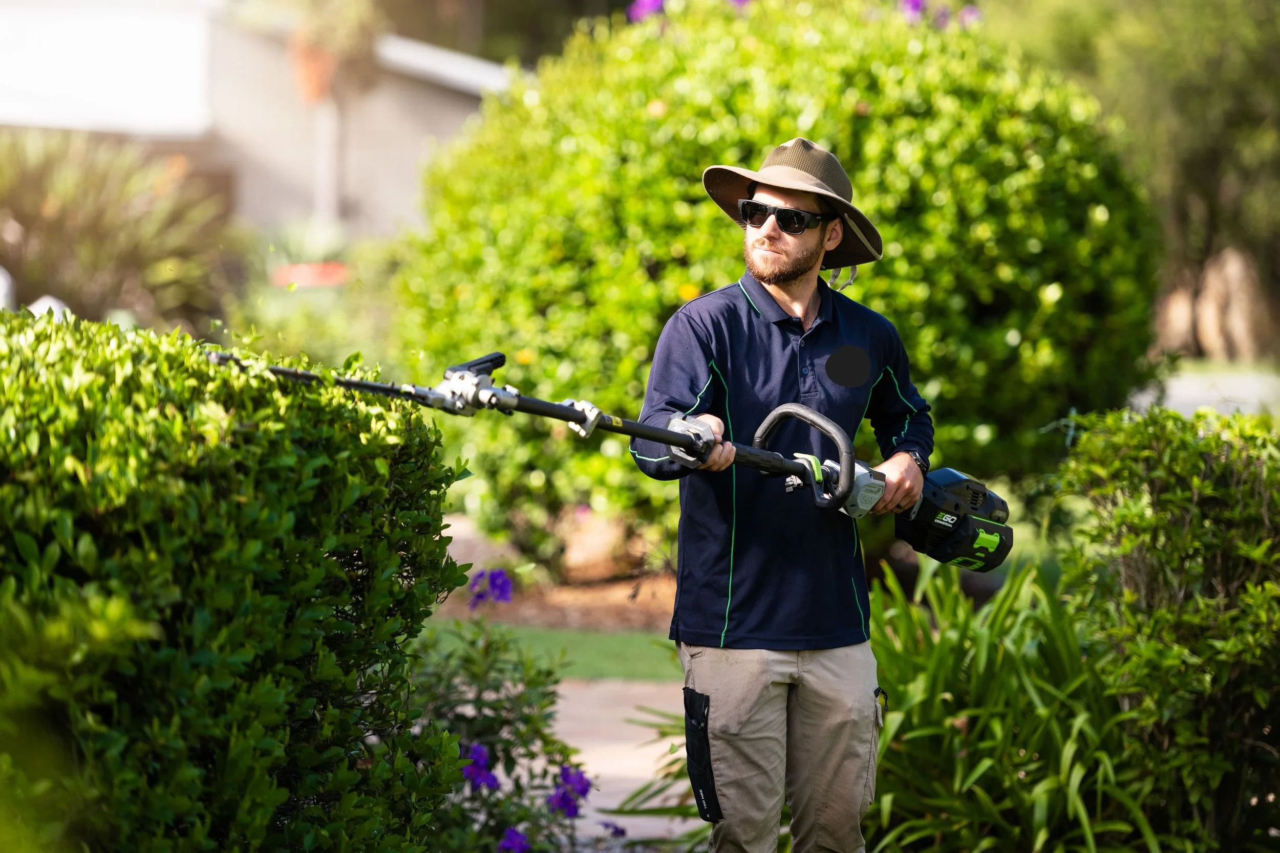 A man wearing a wide-brimmed hat, sunglasses, a navy long-sleeve shirt, and beige cargo pants is trimming bushes with an electric hedge trimmer in a garden filled with green bushes and purple flowers.