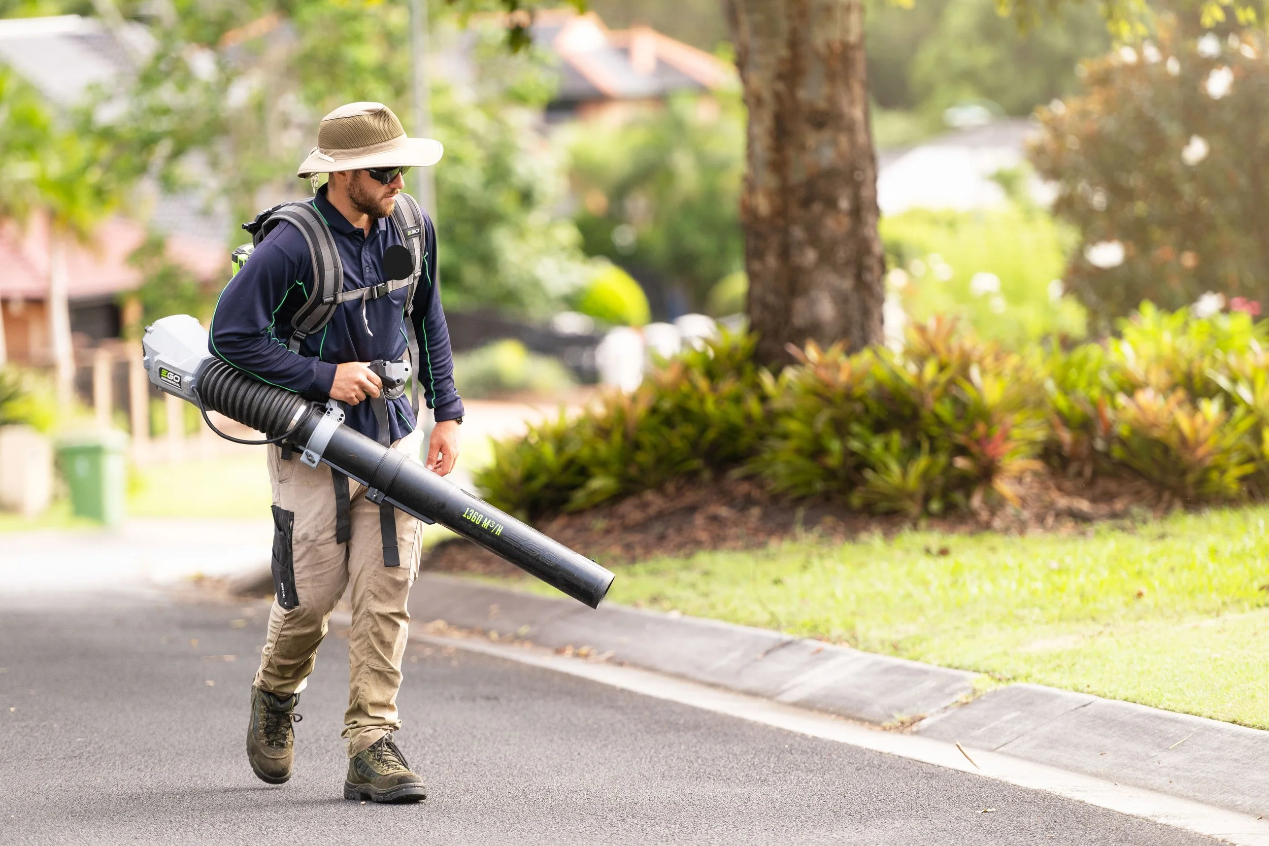 A man outdoors on a suburban street, wearing a wide-brimmed hat, sunglasses, and carrying a leaf blower.