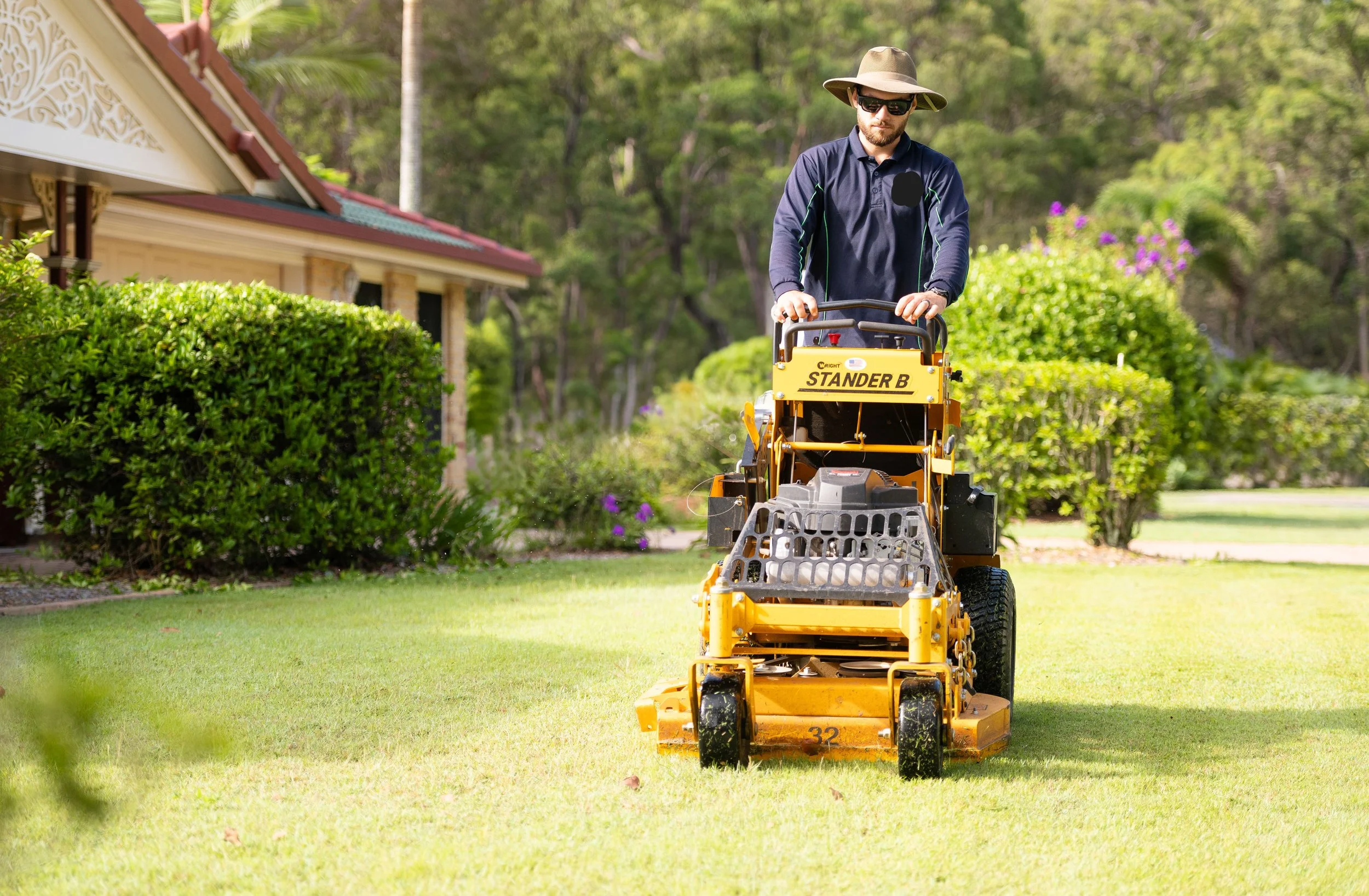 Man wearing sunglasses and a wide-brimmed hat mowing the lawn with a yellow ride-on lawn mower in a garden yard with green bushes and trees.