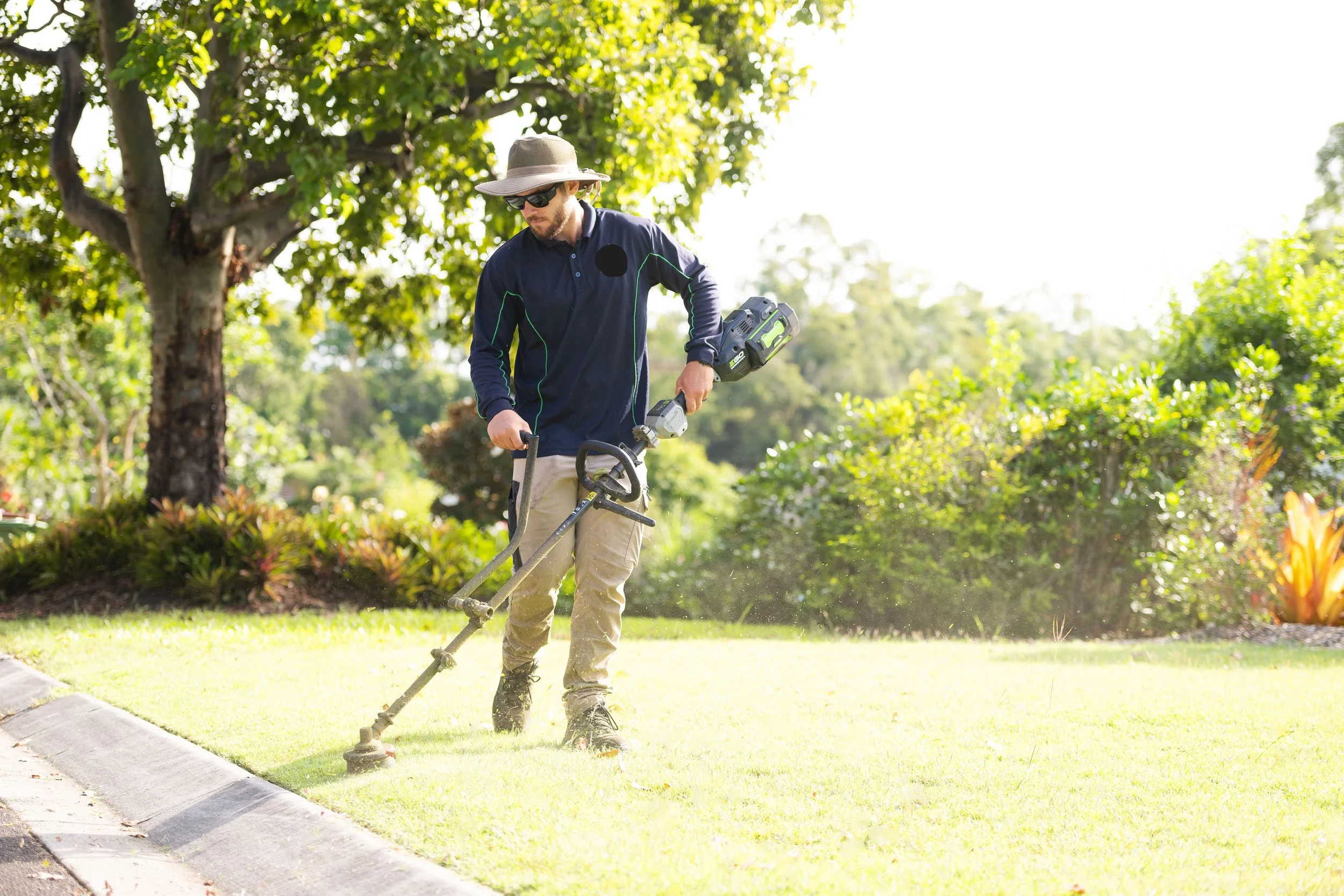 Man wearing sunglasses, a wide-brimmed hat, a navy blue jacket, and khaki pants is using a string trimmer to trim grass along a sidewalk in a park or garden with green shrubs, trees, and sunlight.