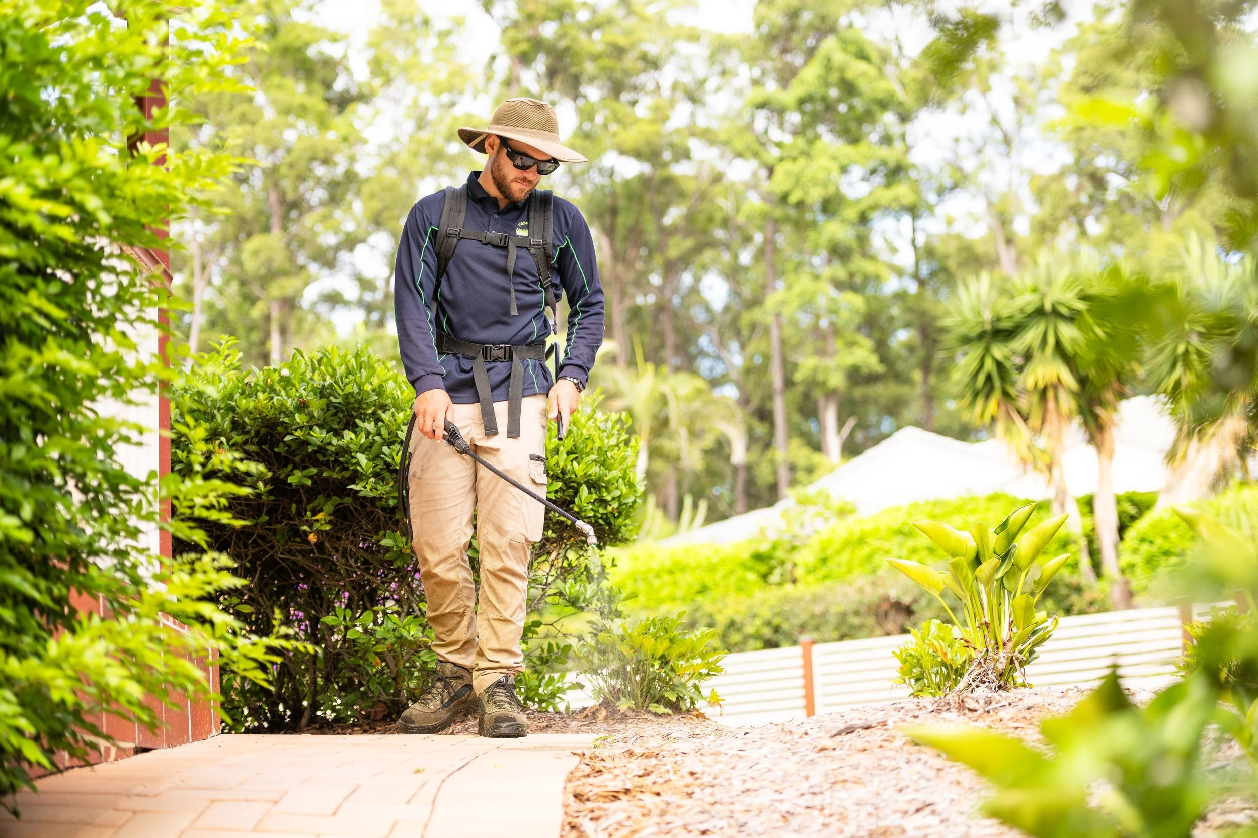 A man in outdoor attire, including a wide-brimmed hat, sunglasses, a navy jacket, and beige pants, is using a gardening tool to spray or weed in a lush garden with green shrubs, trees, and a white fence in the background.